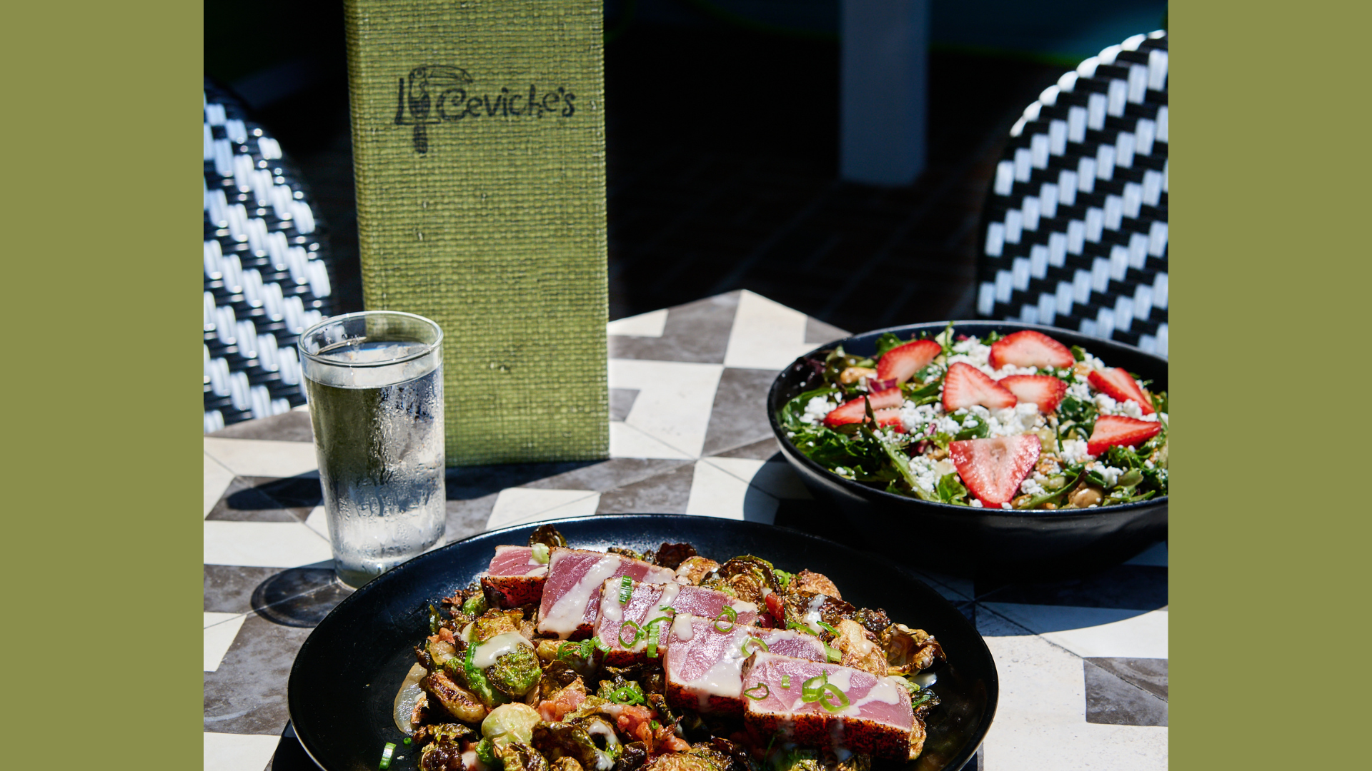 A table outdoors with a glass of water, a bowl of salad with strawberries and cheese, and a plate of cooked meat with vegetables.