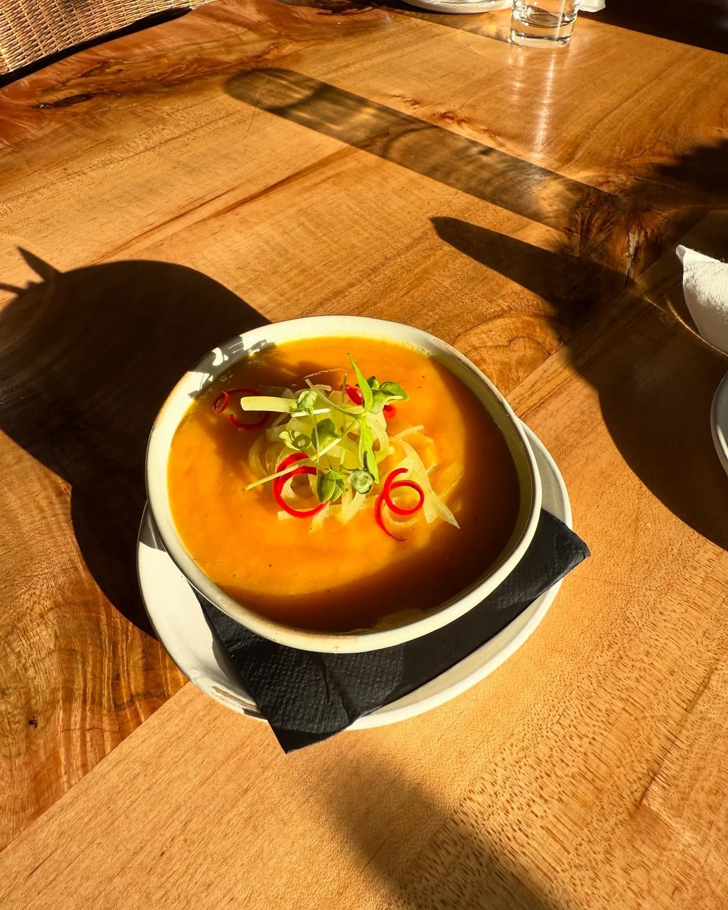 A bowl of orange soup topped with thinly sliced red chili peppers, green microgreens, and sliced onions, placed on a white plate on a wooden table, with a glass of water in the background.