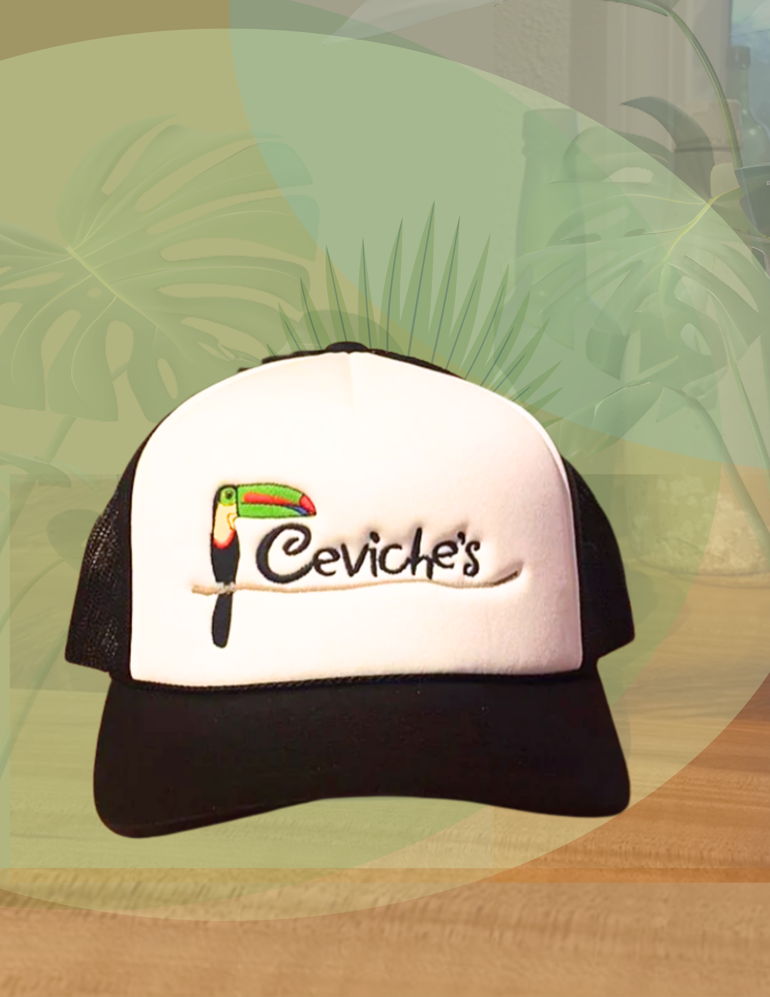 A black and white trucker-style cap with the embroidered logo of a toucan and the word 'Ceviche's' on the front, placed on a wooden surface with a blurred green leafy background.