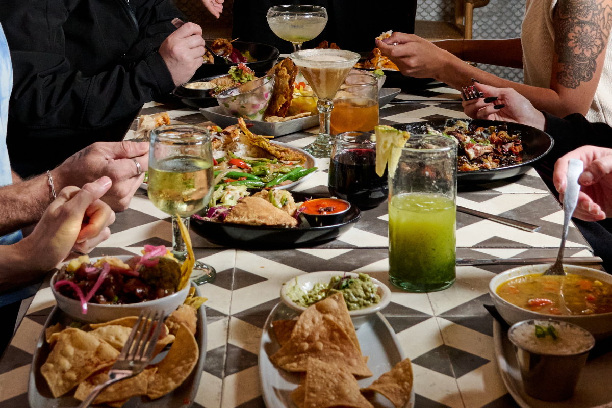 A group of people gathered around a table enjoying a variety of Mexican dishes and drinks, including guacamole, chips, salads, soups, and beverages like margaritas, wine, and green juice.