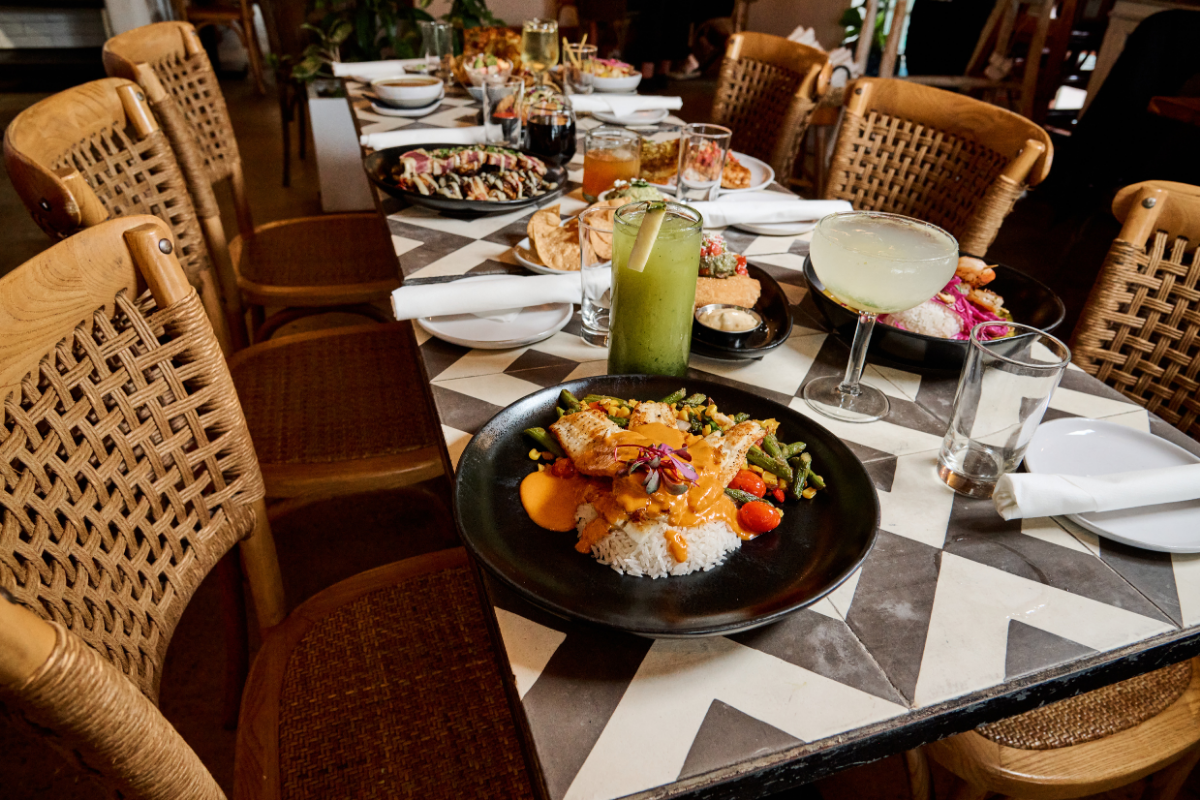 A long dining table set with plates of food, glasses of drinks, and utensils in a restaurant with wooden chairs. The table has a black and white geometric patterned surface.