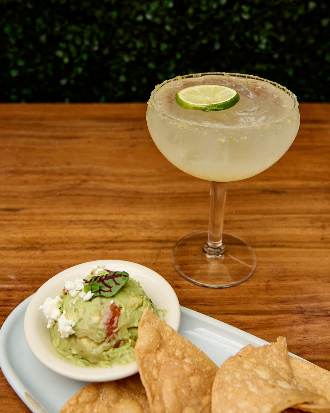 G.O.A.T. Guacamole made with avocado, lime, goat cheese, jalapeños, garlic, pepper, cherry tomato, green onion, topped with micro greens with a side of fresh tortilla chips served on white dishes on a wood bar, next to a Sea Love Margarita featuring 