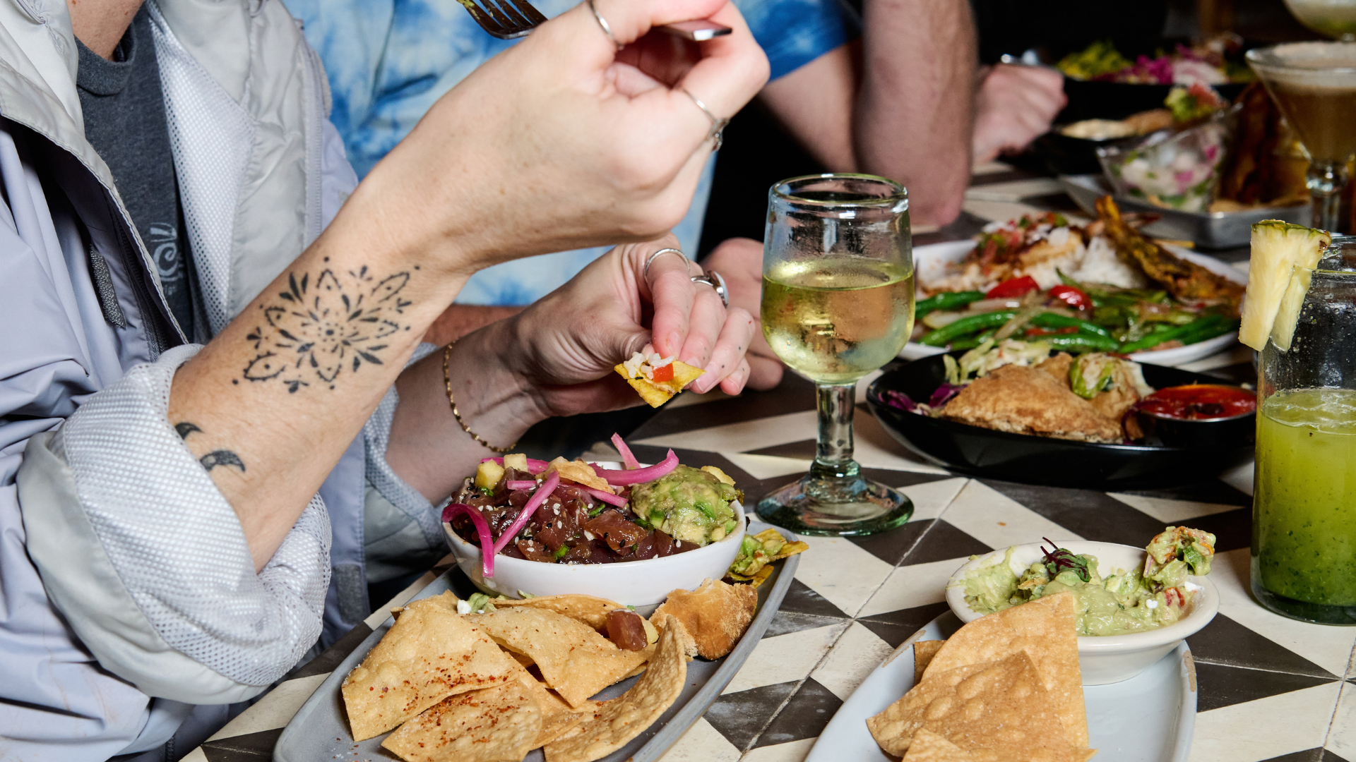 People sharing a variety of Mexican dishes on a table, including chips, guacamole, salsas, and drinks.