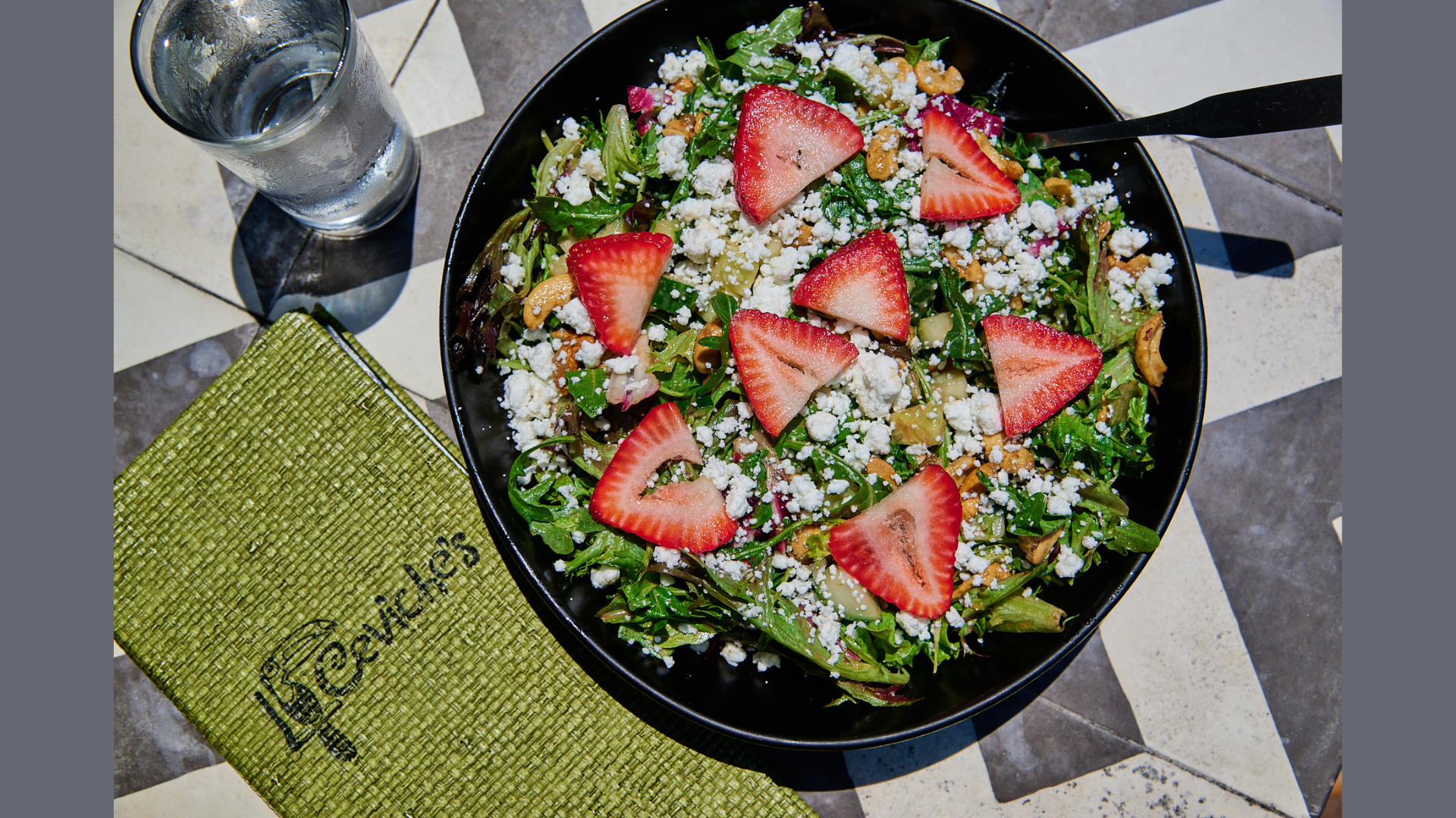 A salad with mixed greens, sliced strawberries, crumbled feta cheese, and nuts in a black bowl on a tiled table, accompanied by a glass of water and a green napkin.