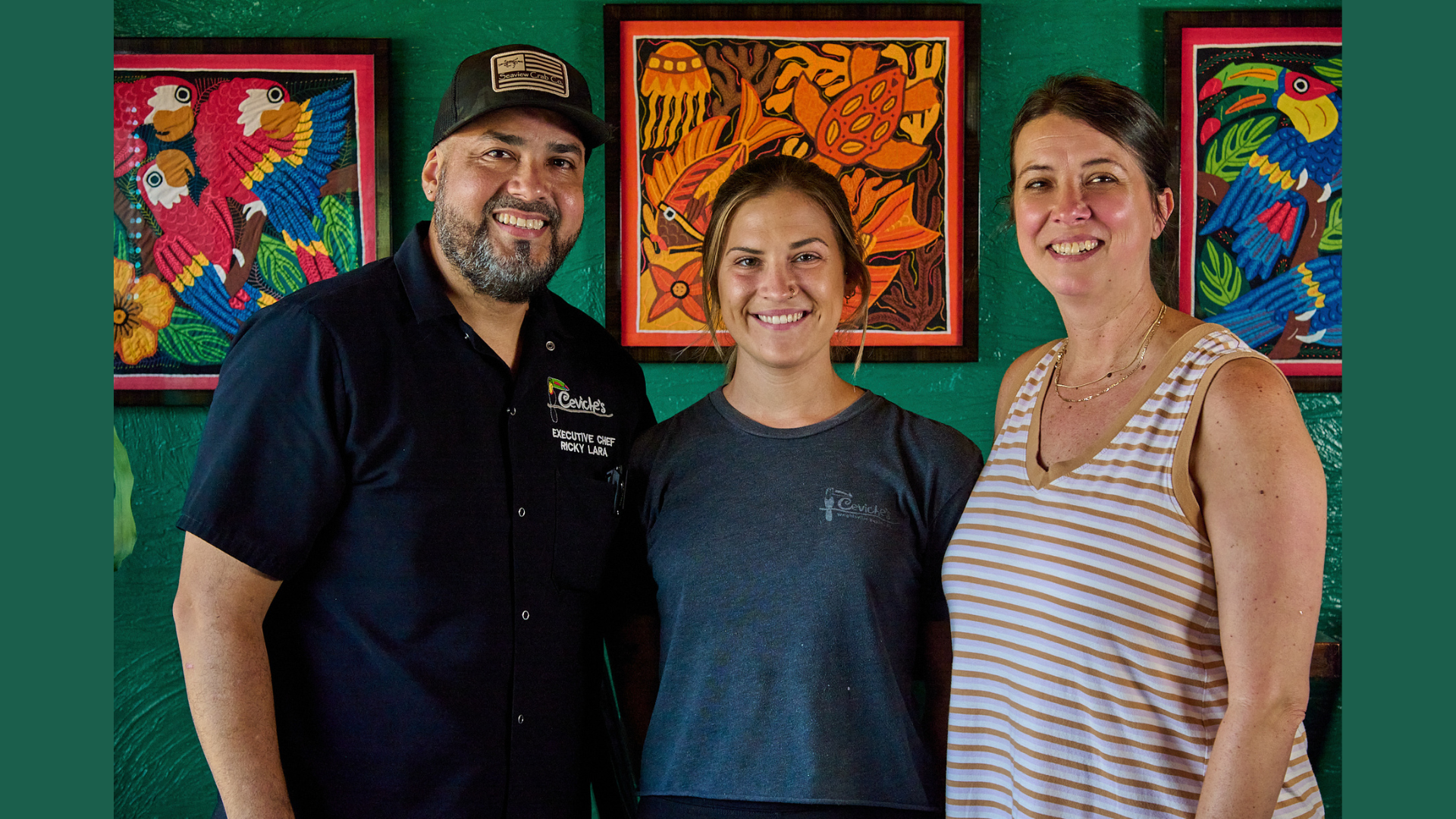 Three people smiling inside an art gallery, standing in front of colorful framed artwork featuring tropical birds and flowers.