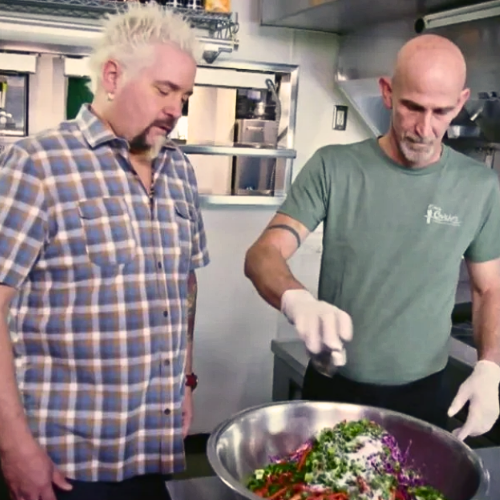 Two men preparing food in a kitchen, one is wearing a blue plaid shirt and the other a green T-shirt, handling vegetables in a large bowl.