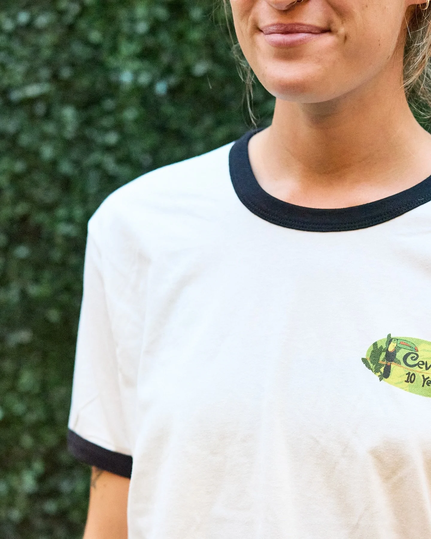 Close-up of a woman wearing a white T-shirt with black collar and sleeve trim, standing outdoors with greenery in the background.