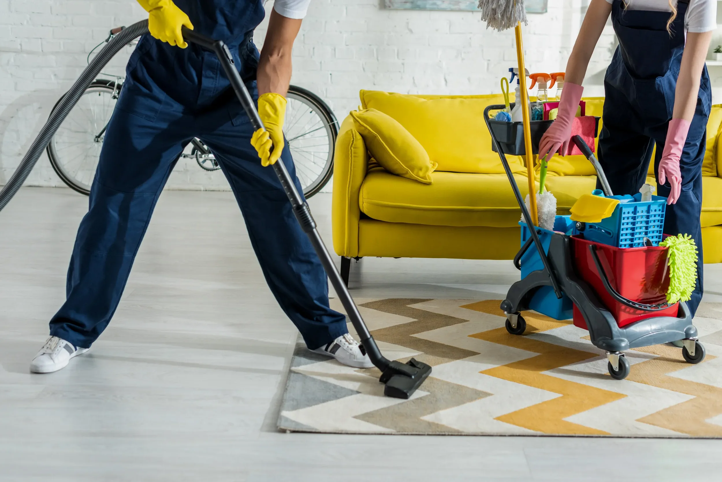 Cleaning service near Perth Australia. Two people cleaning a living room with multiple cleaning supplies, one wearing yellow gloves and vacuuming the carpet, the other reaching into a cleaning cart, with a yellow sofa and bicycle in the background.