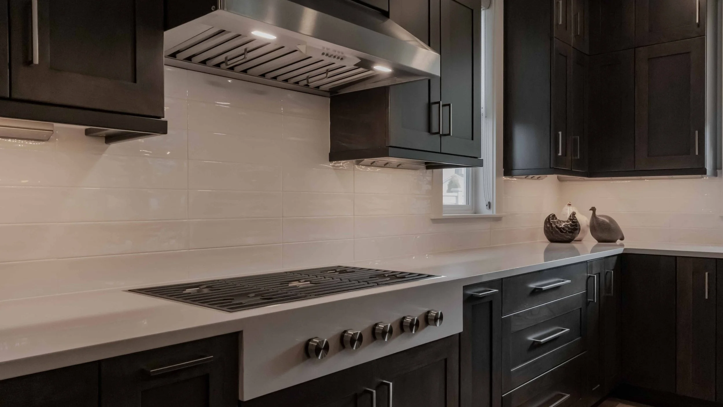 Modern kitchen with black cabinetry, white countertops, built-in stove, and decorative ceramic chicken and hen figurines on the counter.