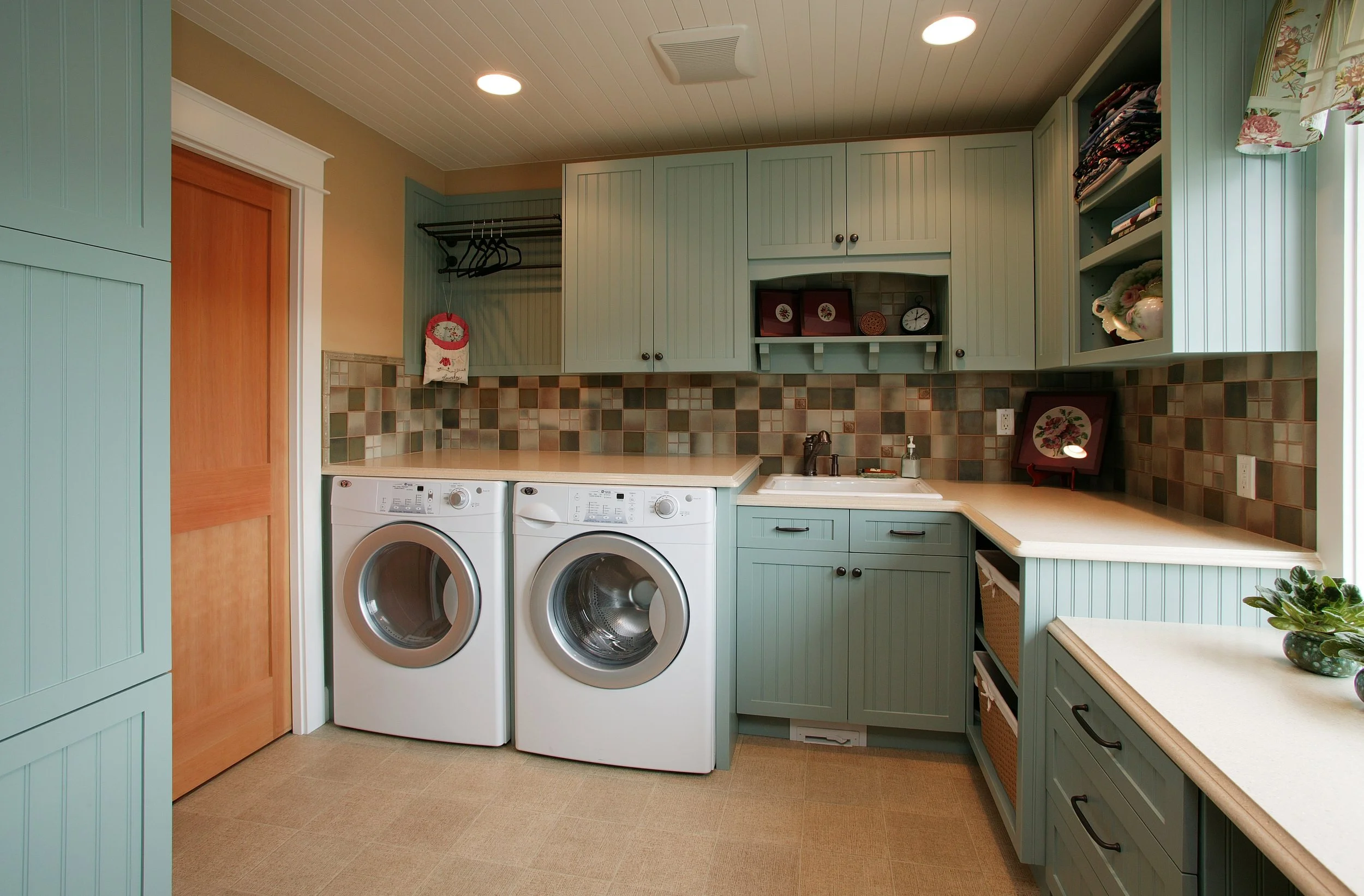 Laundry room with teal cabinets, a washer and dryer, beige countertop, spray soap on the sink, tiled backsplash, and a window with floral curtains.