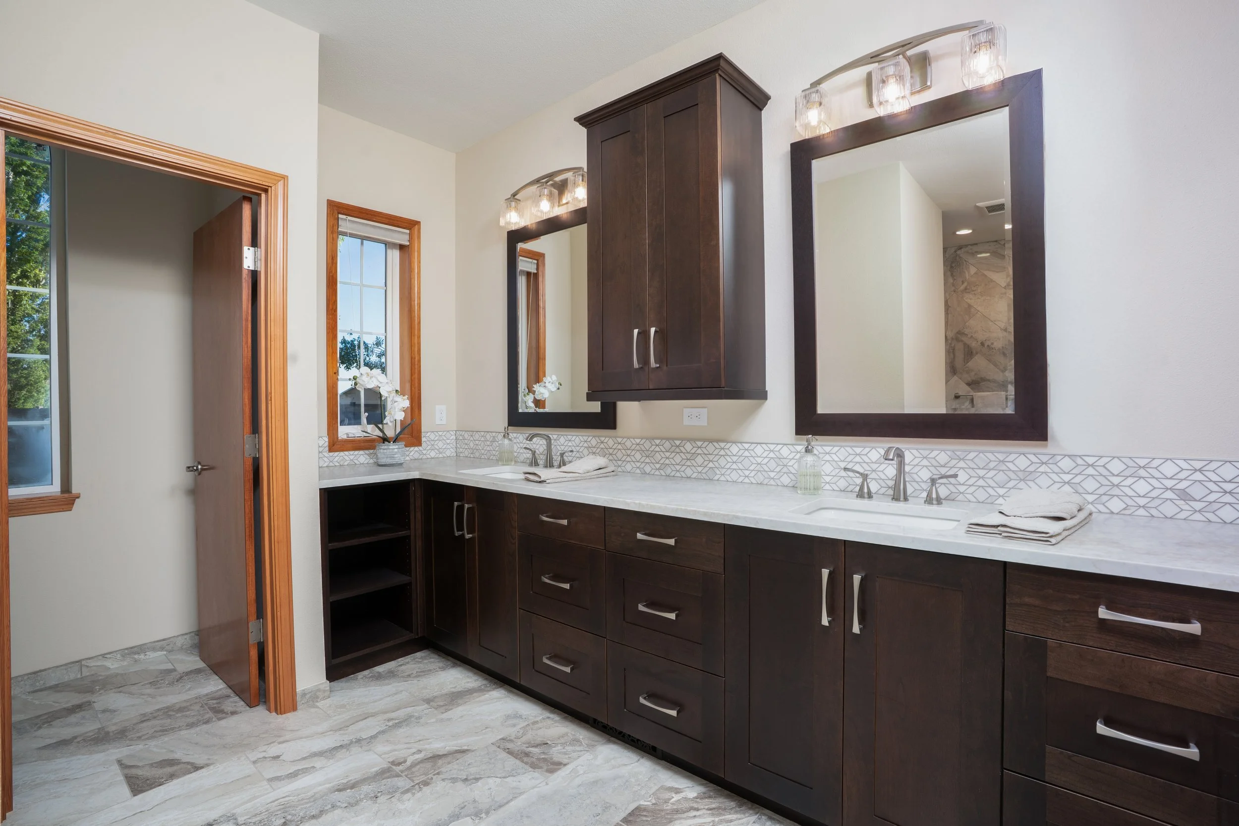 Remodeled bathroom with a dark wood double vanity, marble-look counters, and gray tile floor by Choice Carpentry