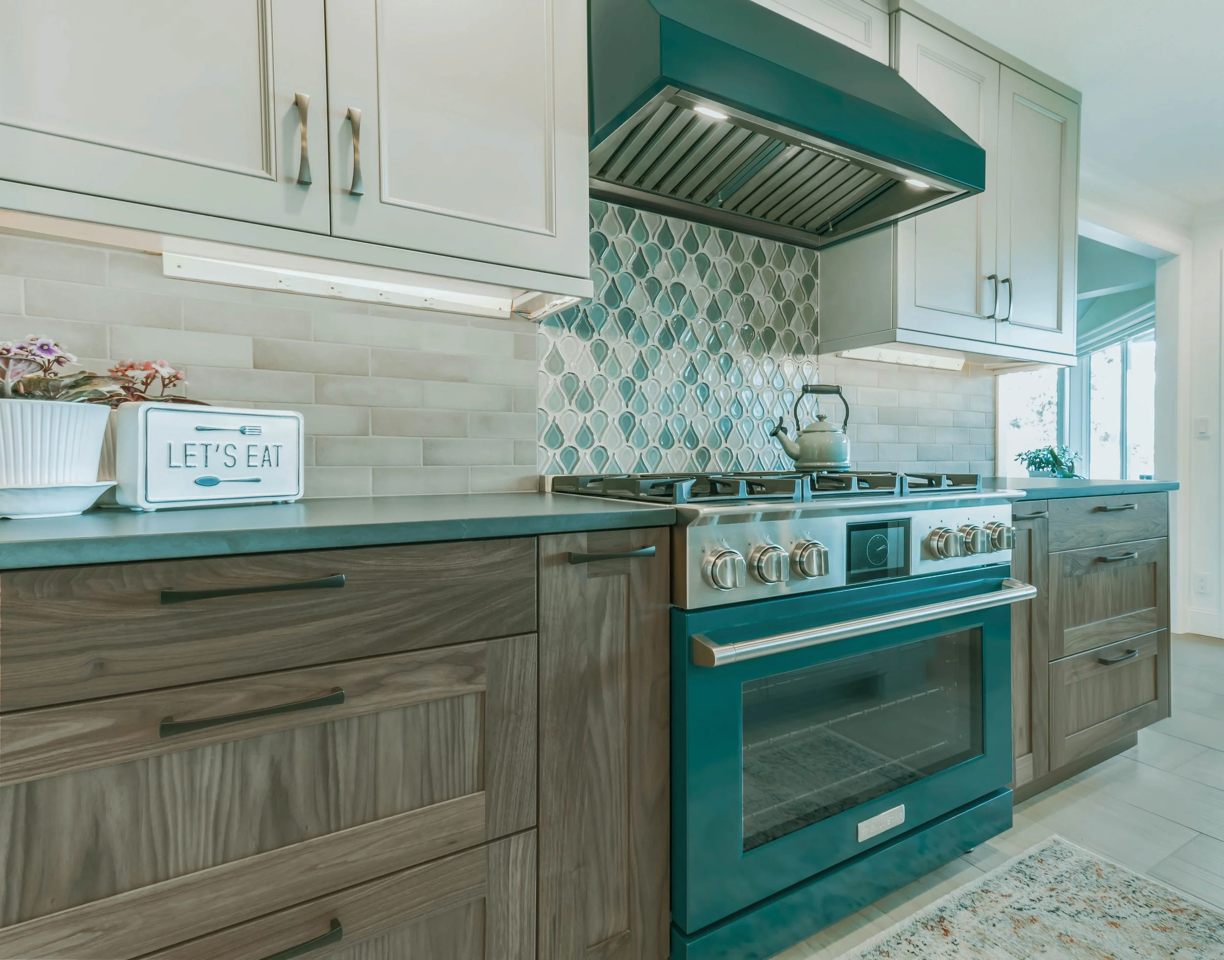 A modern kitchen with a teal gas stove, beige cabinetry, a decorative tile backsplash, and a window with plants.