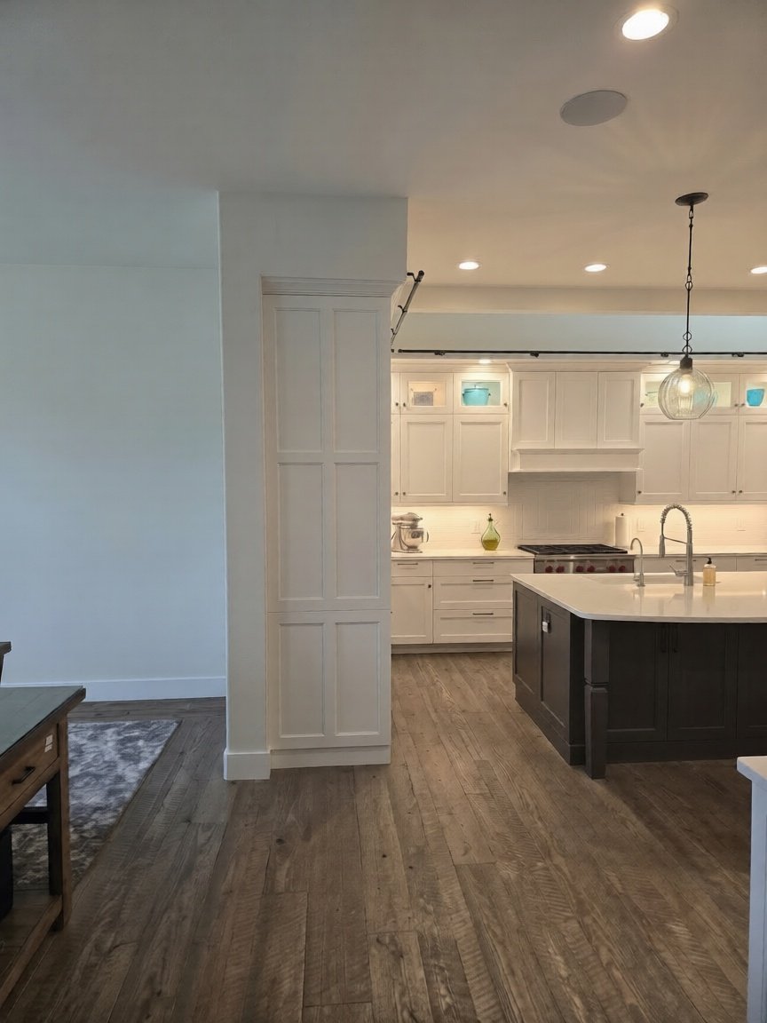 Remodeled kitchen seen through a doorway, with a dark island, white cabinetry, glass pendant lights and wood-look flooring, by Choice Carpentry