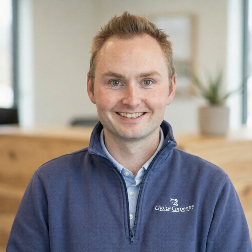 A smiling man in a blue zip-up jacket with 'Choice Carpentry' logo stands in front of a wall covered with architectural blueprints.