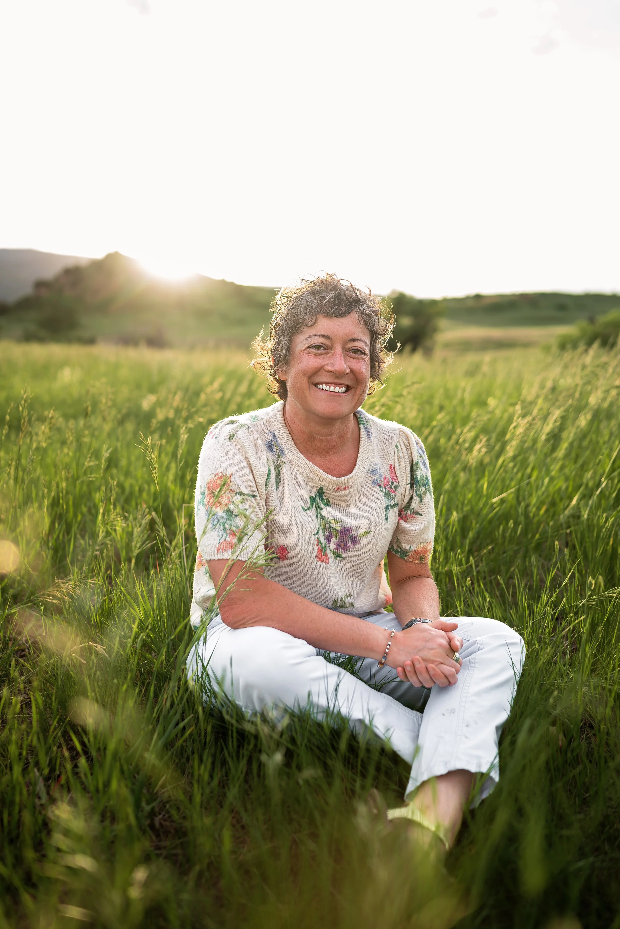 A woman with curly hair sits on grass in a field at sunset, smiling at the camera, wearing a floral sweater and white pants.