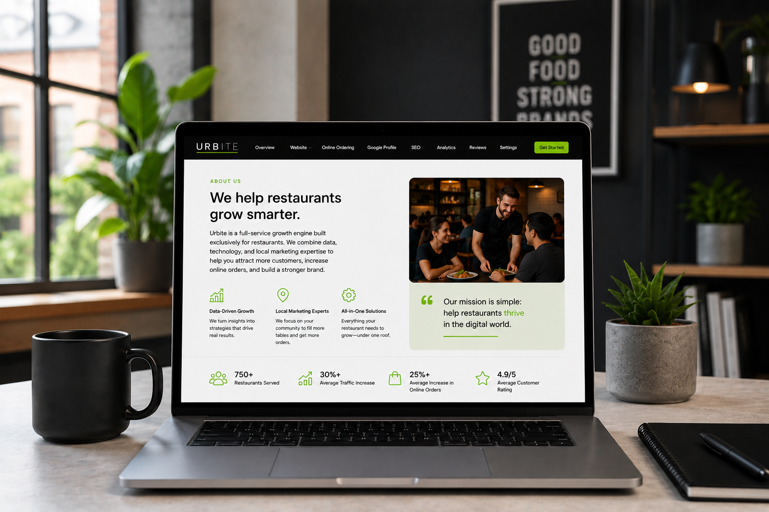 Open laptop on a desk displaying a website for Urbite, a company that helps restaurants grow through digital. The page features text, icons, and a photo of a smiling waiter serving food to customers in a restaurant. There are two potted plants and a black coffee mug on either side of the laptop, and a notebook with a pen to the right.
