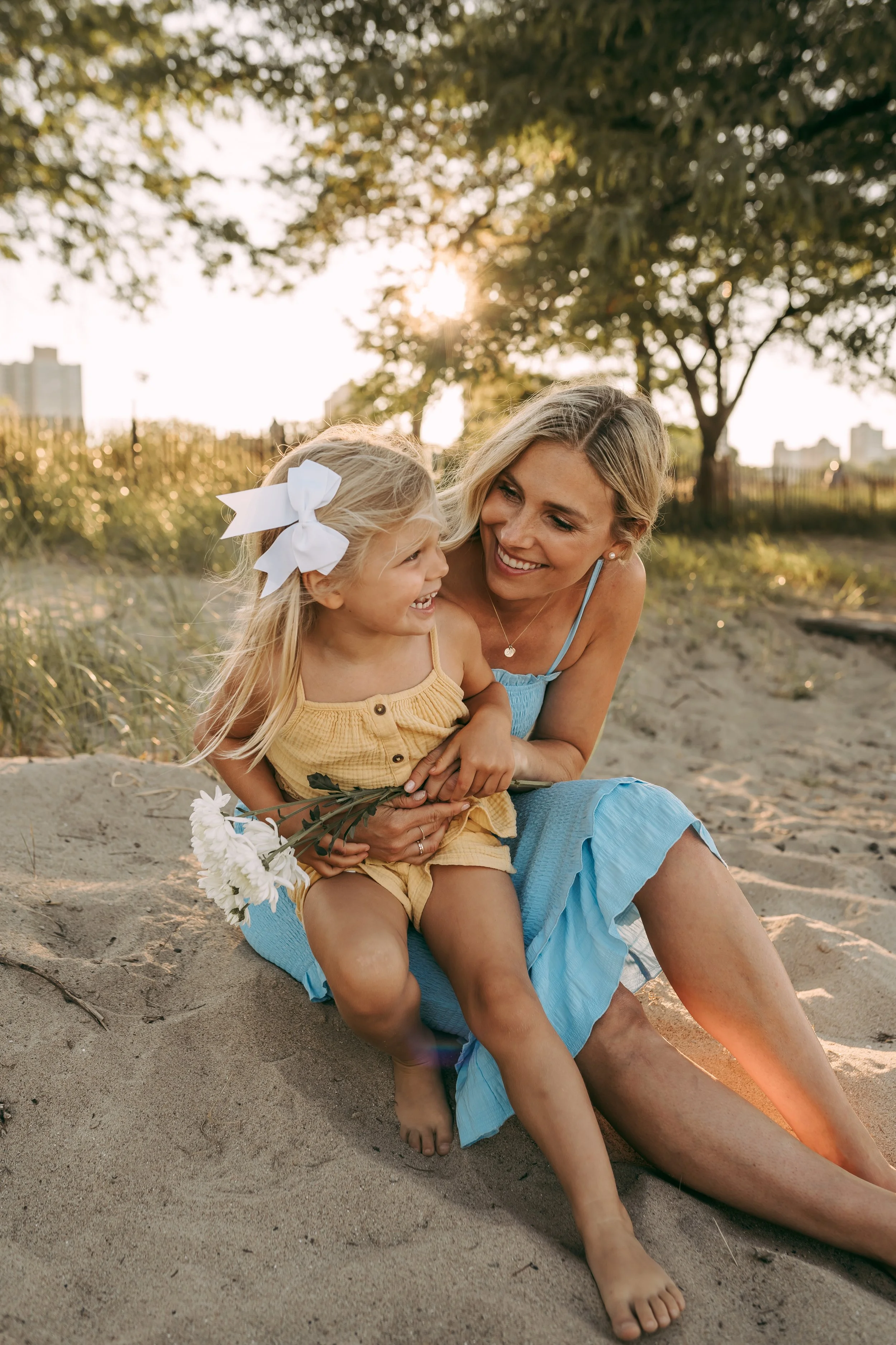 A woman and a young girl sitting on the sand at a beach, smiling and enjoying the sunset, with trees and city buildings in the background.