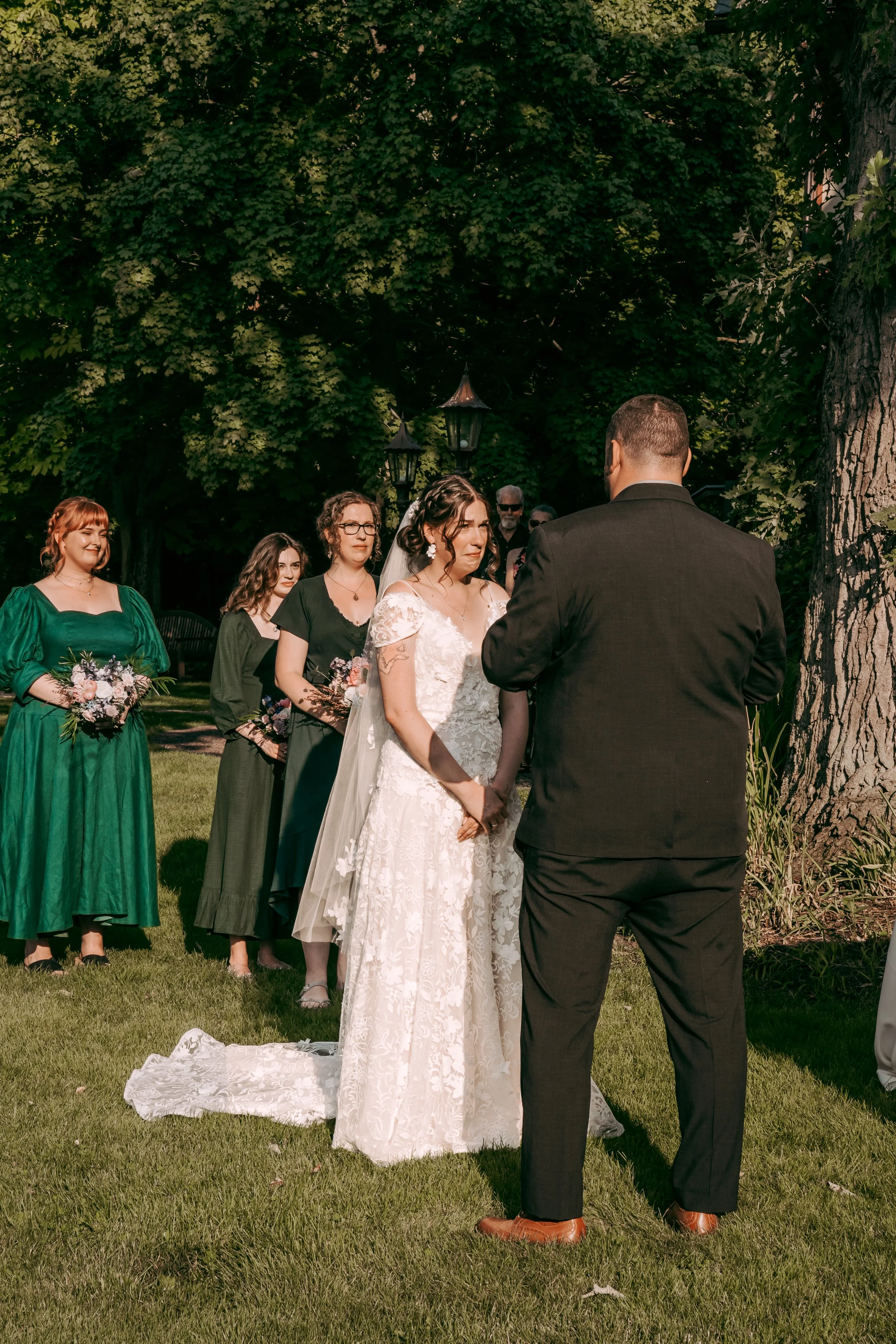 A bride and groom exchanging vows outdoors during a wedding ceremony, with bridesmaids standing in a row behind the bride and groom, on a sunny day with green trees in the background.