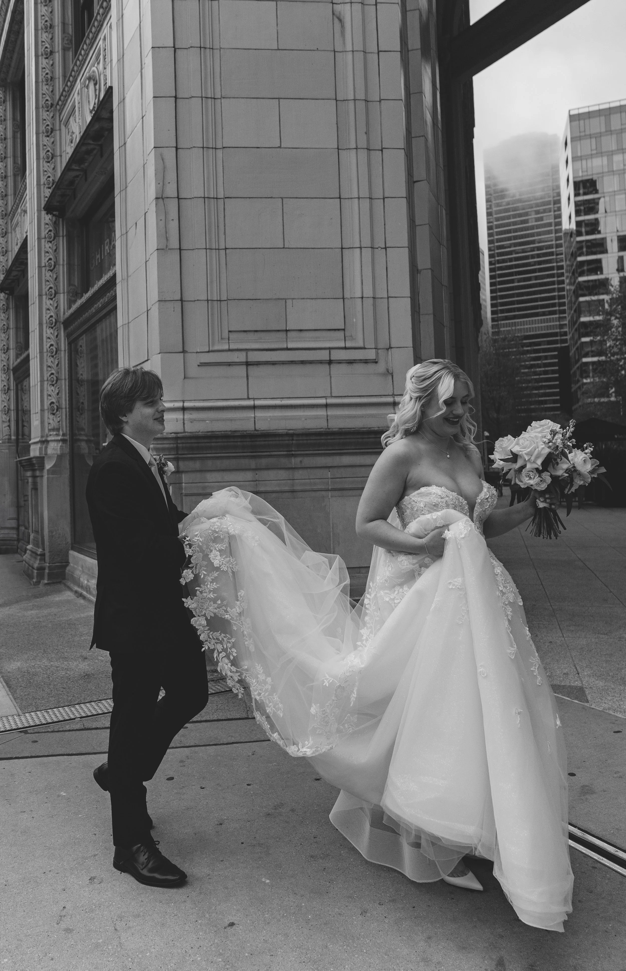 Black and white photo of a bride in a wedding dress holding a bouquet, assisted by a groom in a suit, outside a tall building in an urban setting.