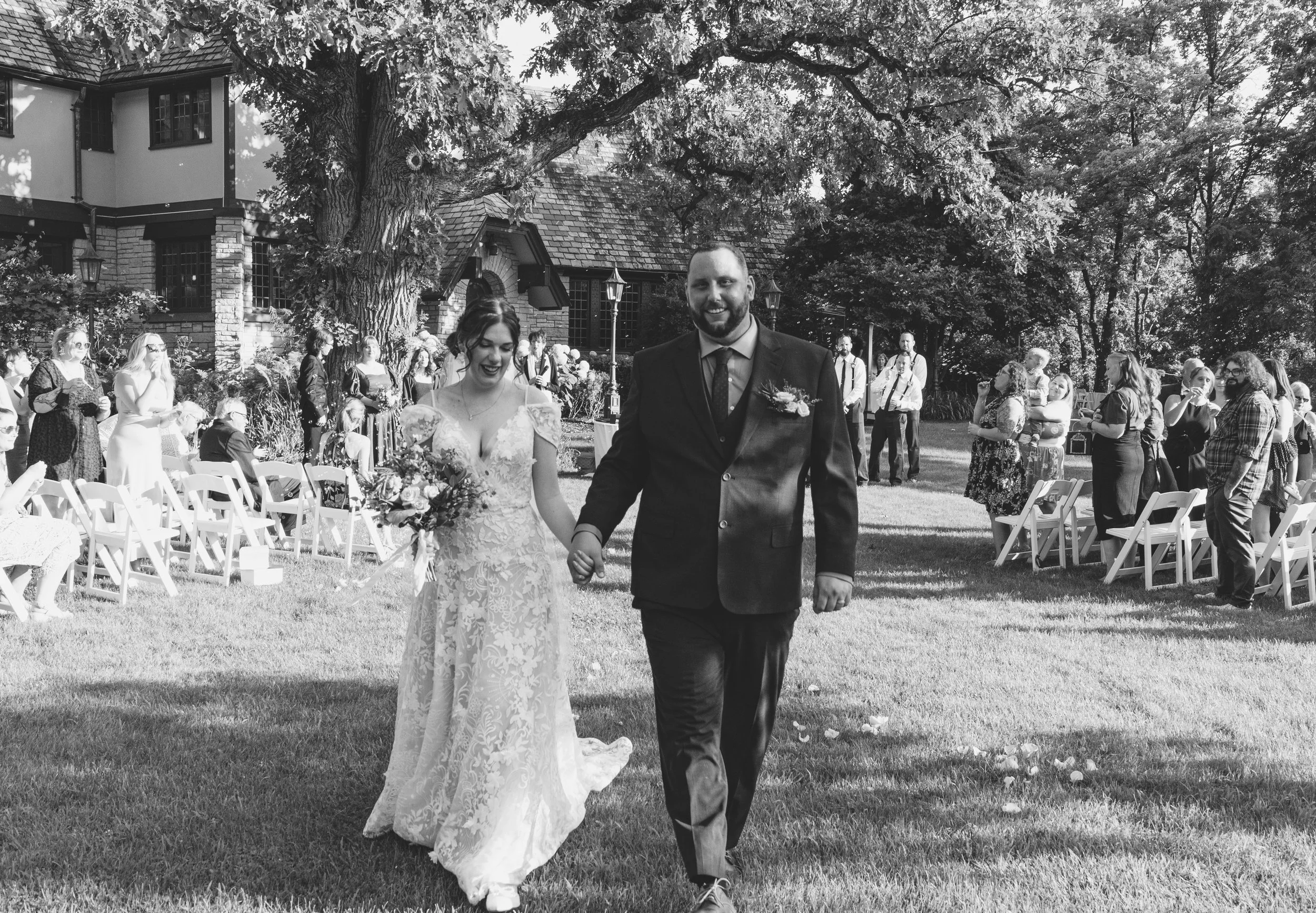 A couple in wedding attire walking hand-in-hand outdoors, surrounded by seated and standing wedding guests, in a garden area with large trees and a house in the background