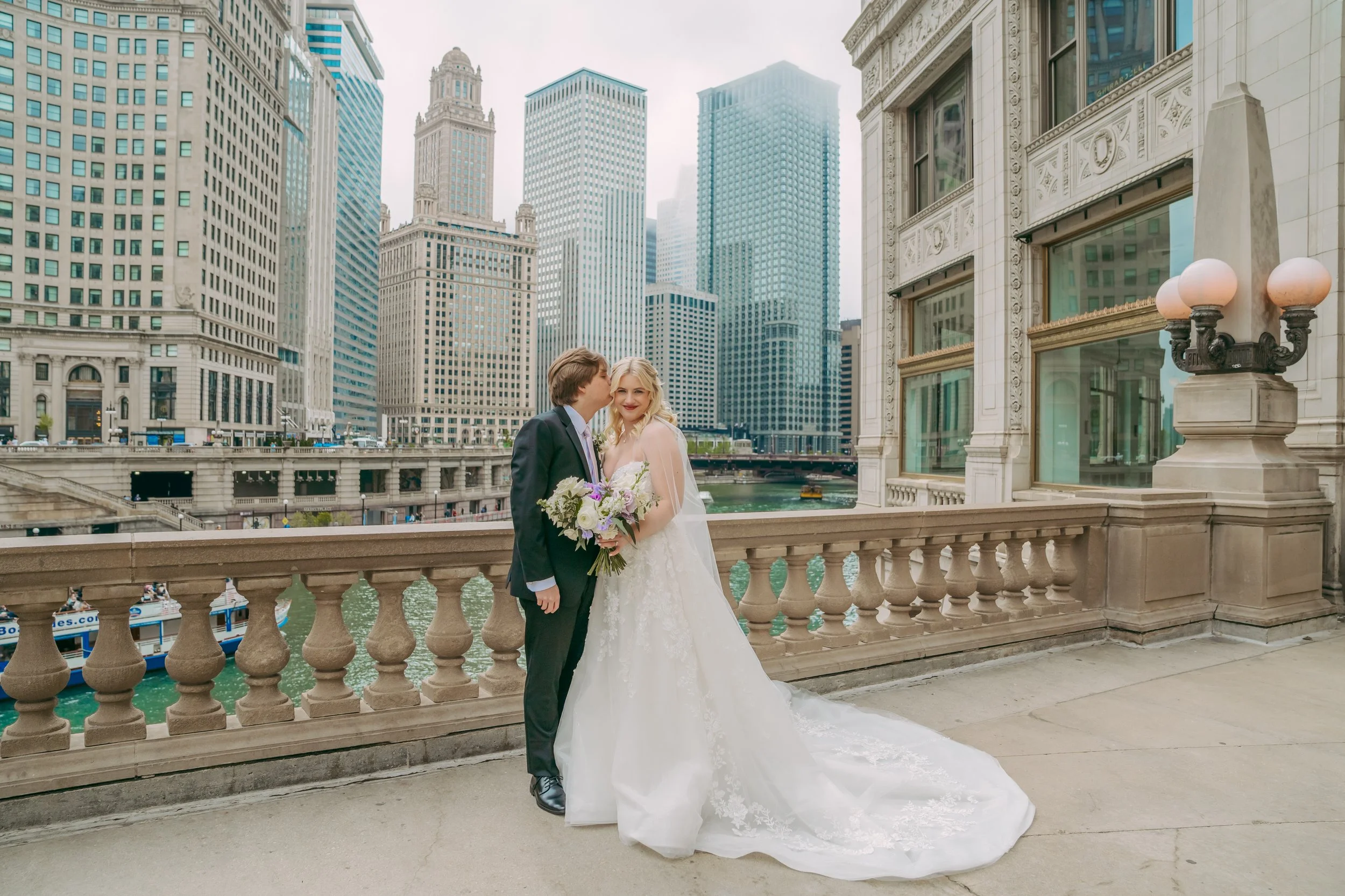 A bride and groom pose for a wedding photo on a balcony with a city skyline of tall buildings in the background.