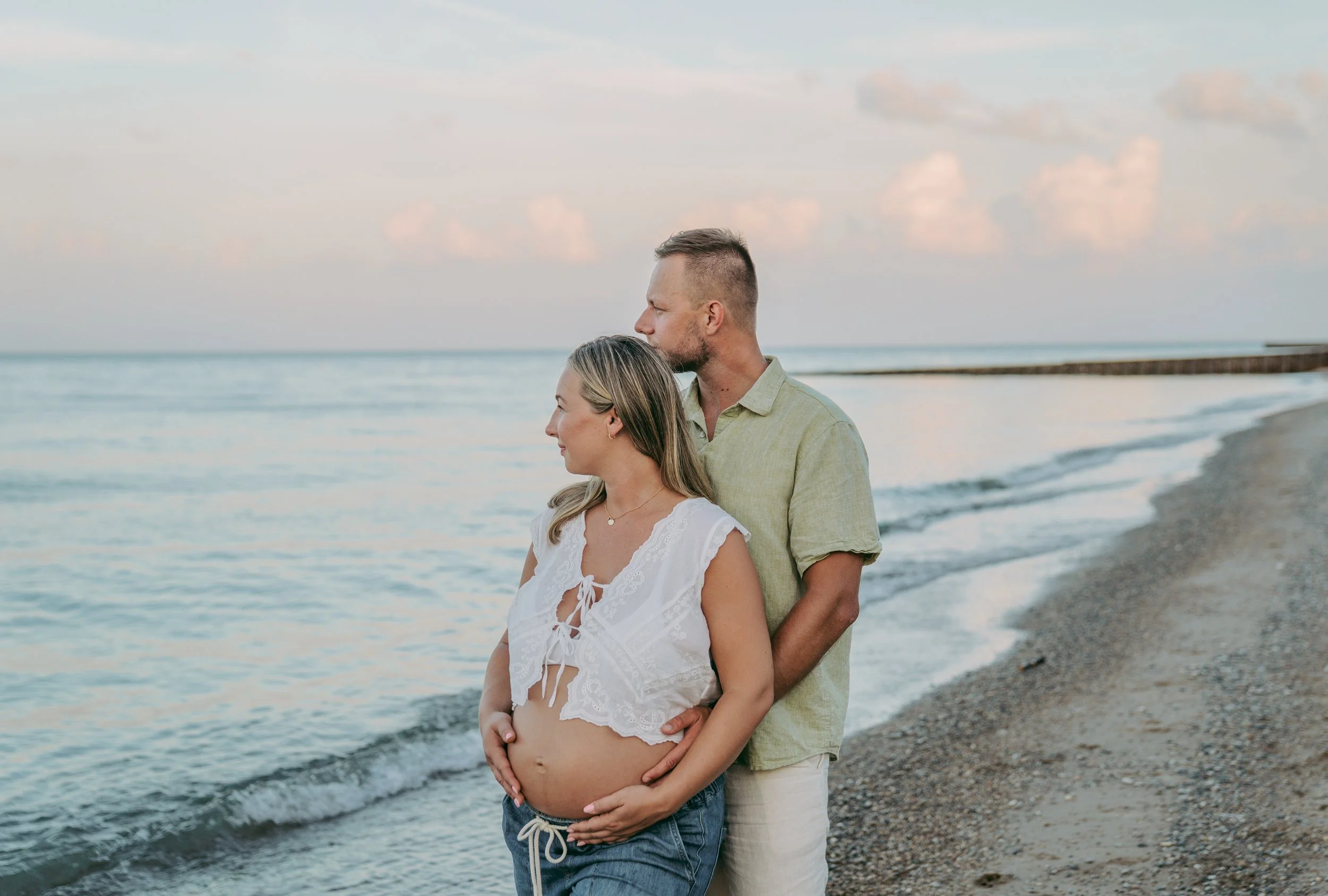 A pregnant woman and a man standing on a beach with the ocean in the background. The woman has her hands on her belly, and the man is kissing her head.