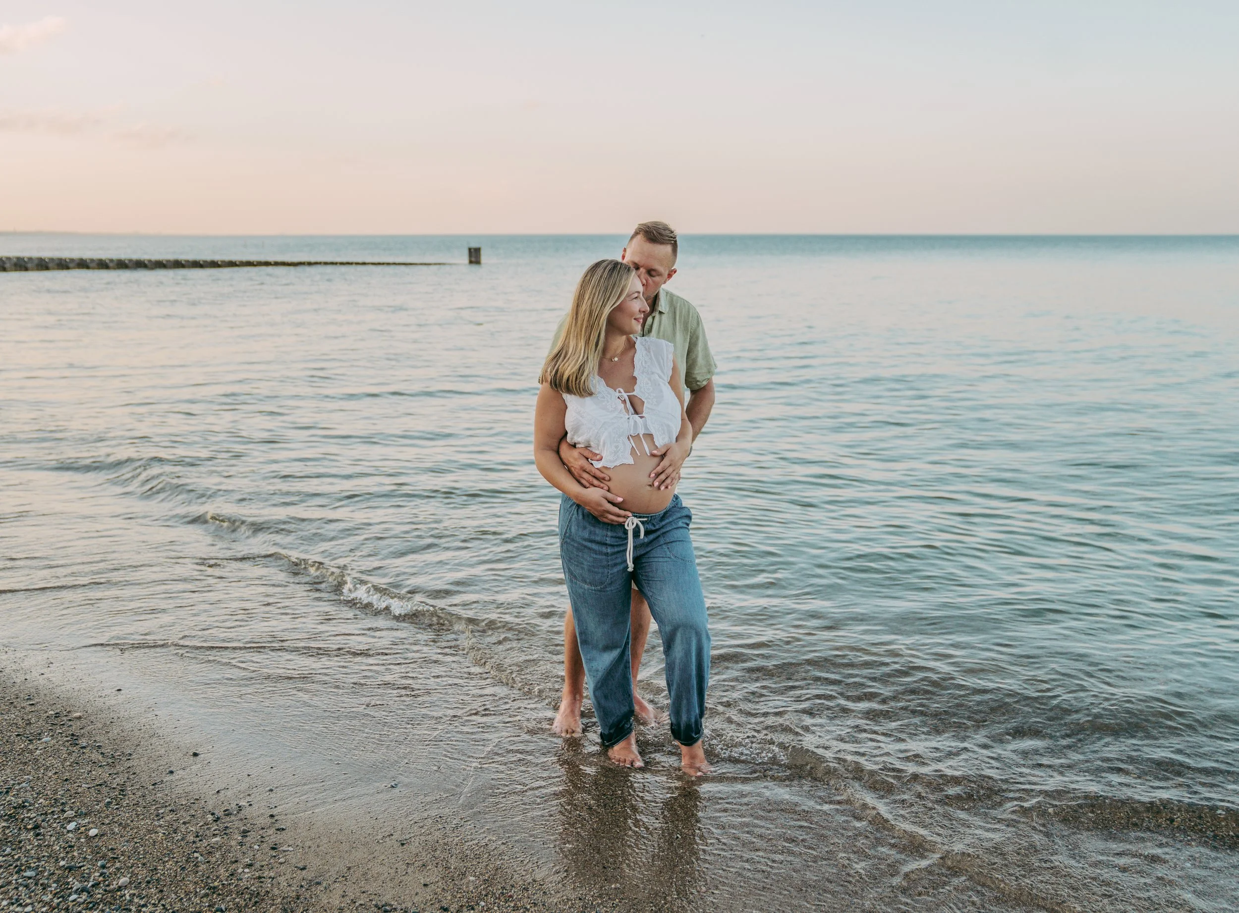 Pregnant woman and man standing in shallow water at the beach during sunset, with the woman holding her belly and the man embracing her from behind.
