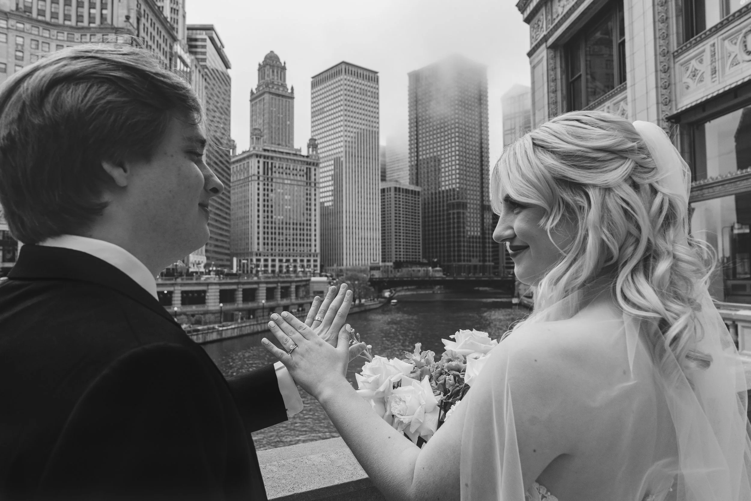 Black and white photo of a bride and groom touching forearms on a city bridge with skyscrapers in the background.
