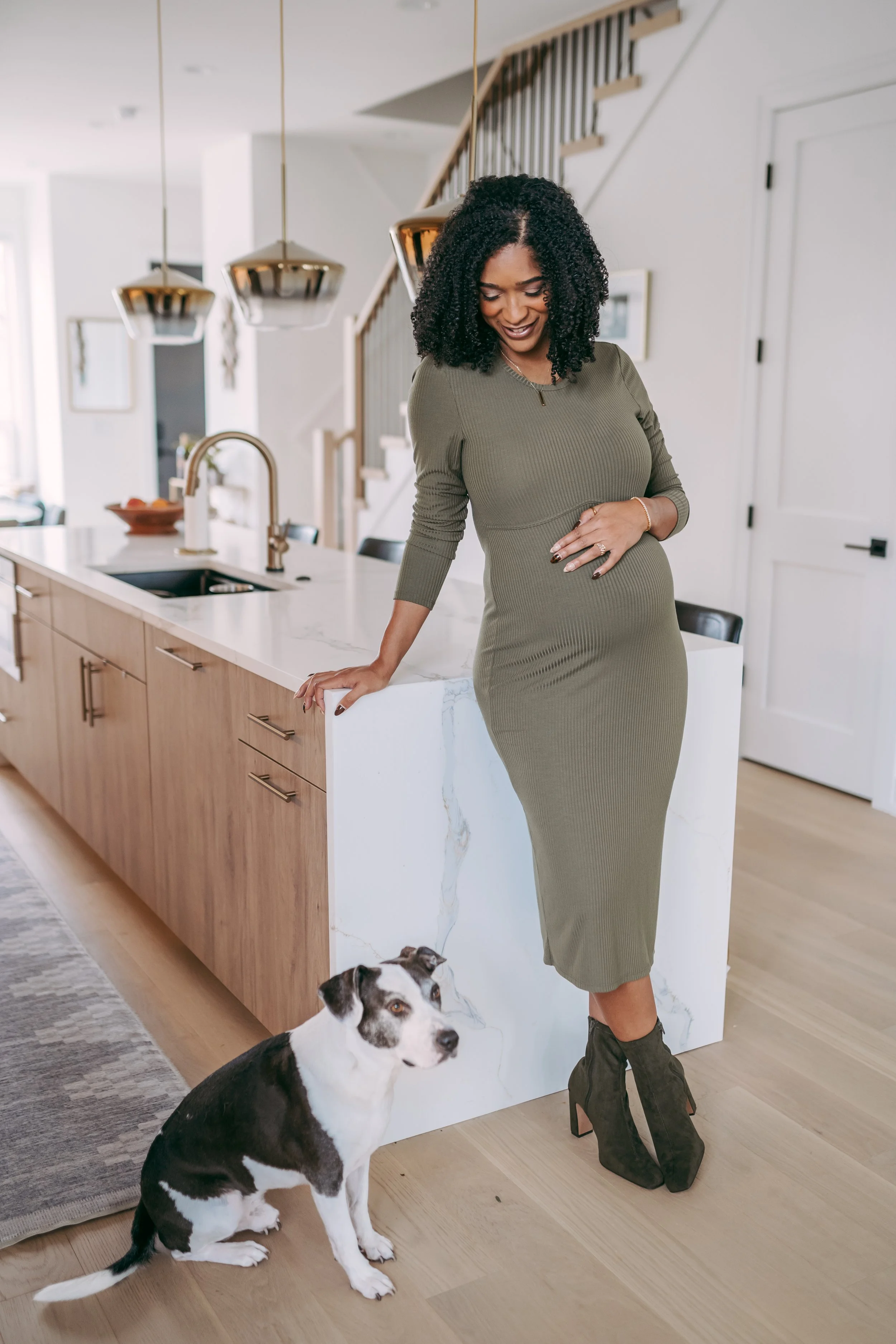 A woman with curly black hair in a fitted olive green dress and high heeled shoes standing in a modern kitchen with a black and white dog sitting beside her, on a light wooden floor, with a white marble kitchen countertop and wooden cabinets.