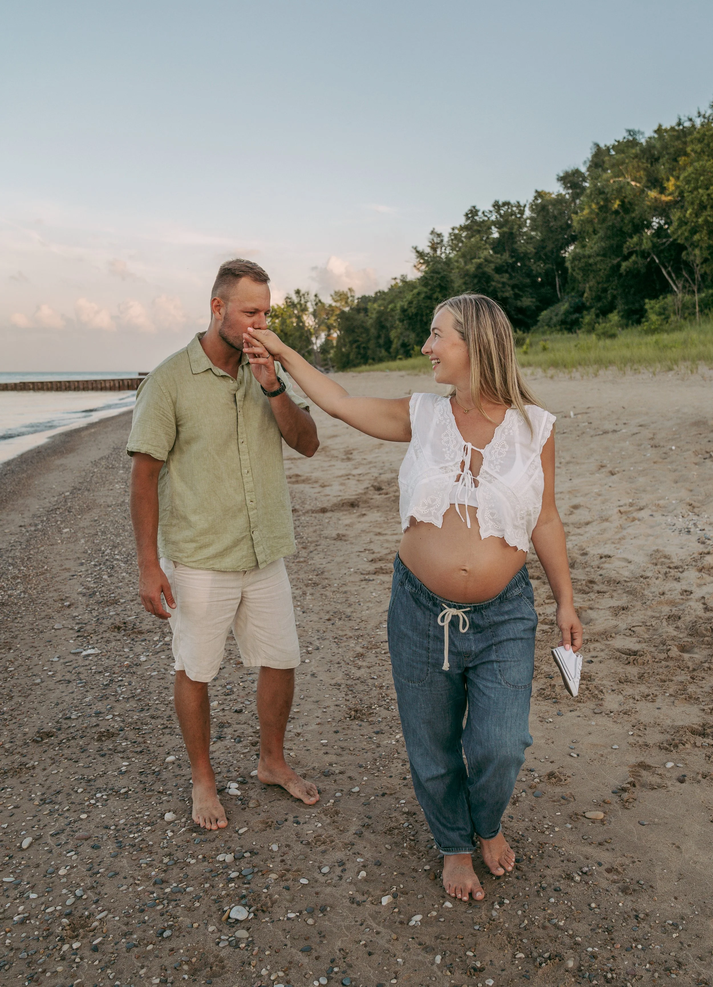 A pregnant woman on the beach touching her partner's face, who is kissing her hand, both smiling, during sunset.