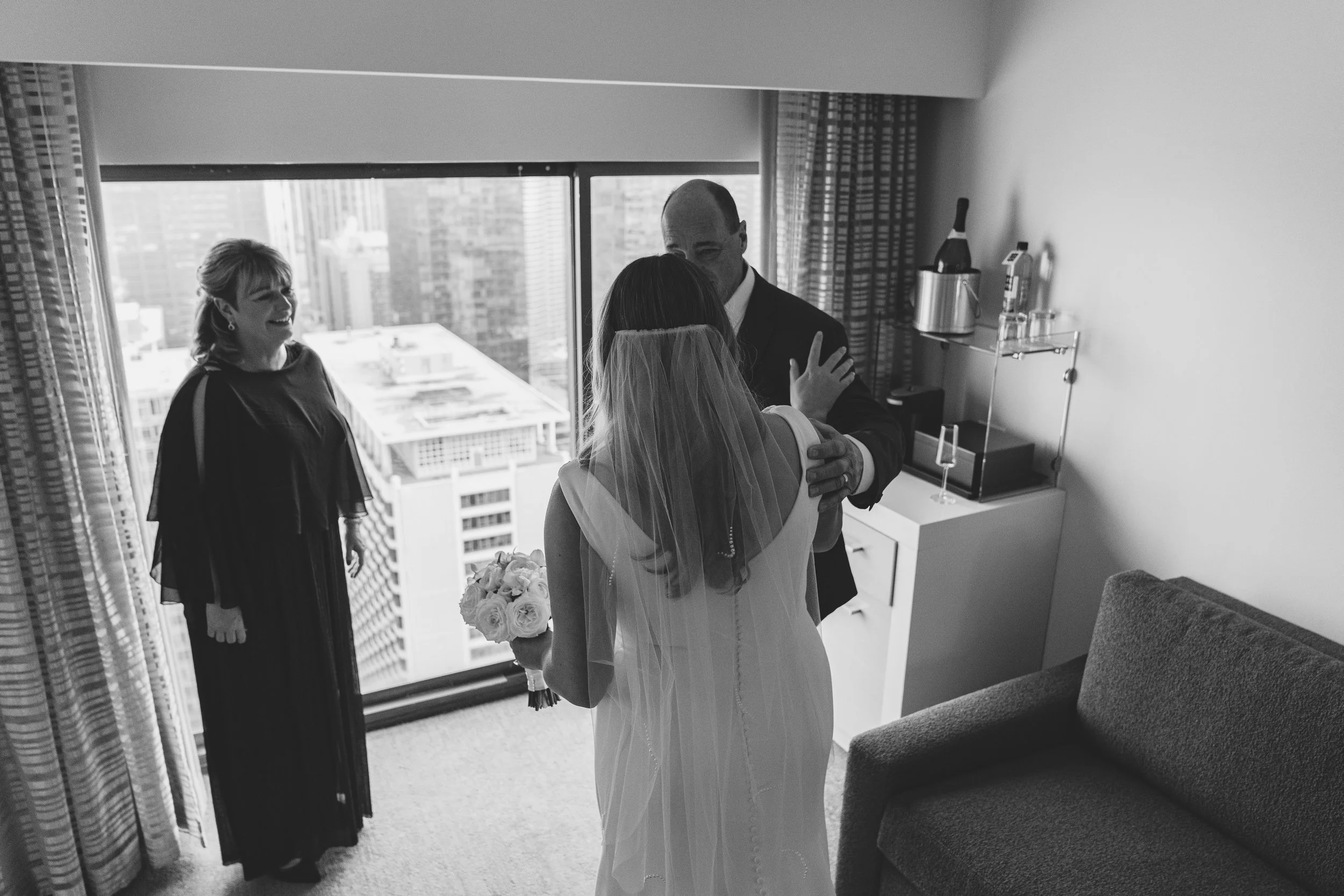 A bride hugging a man, likely her father, while a woman looks on in a hotel room with cityscape view in the background.