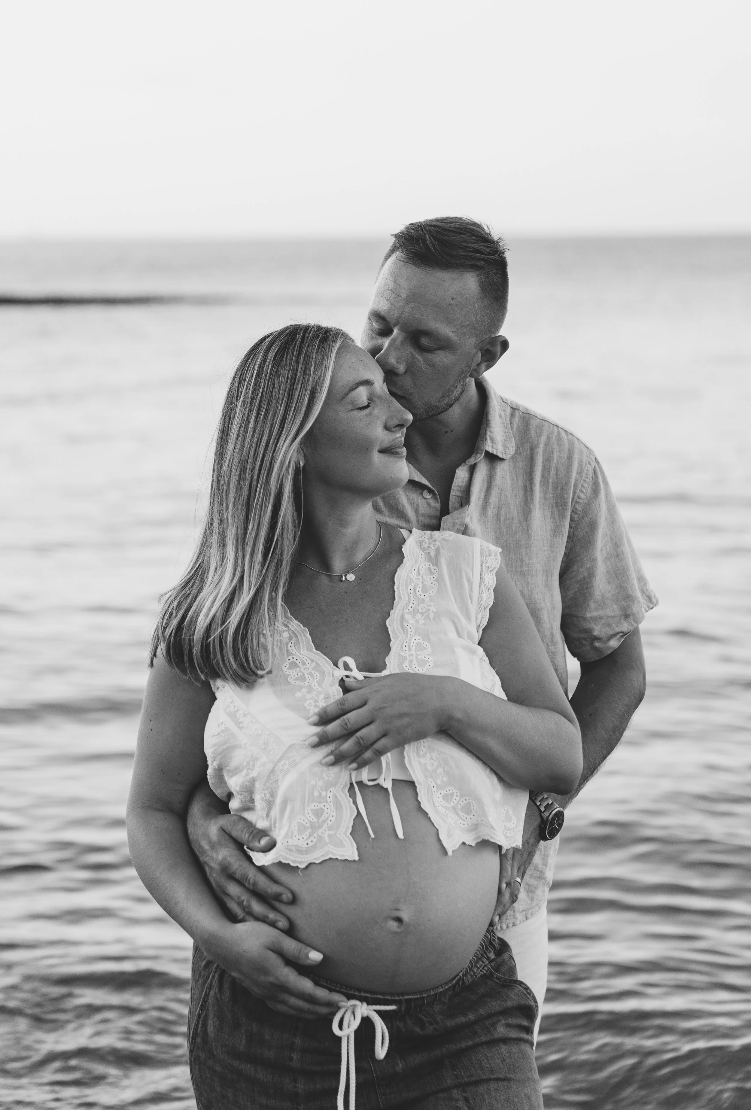 A black and white photo of a pregnant woman and a man standing by the water. The man is kissing the woman's forehead while she gently touches her belly.