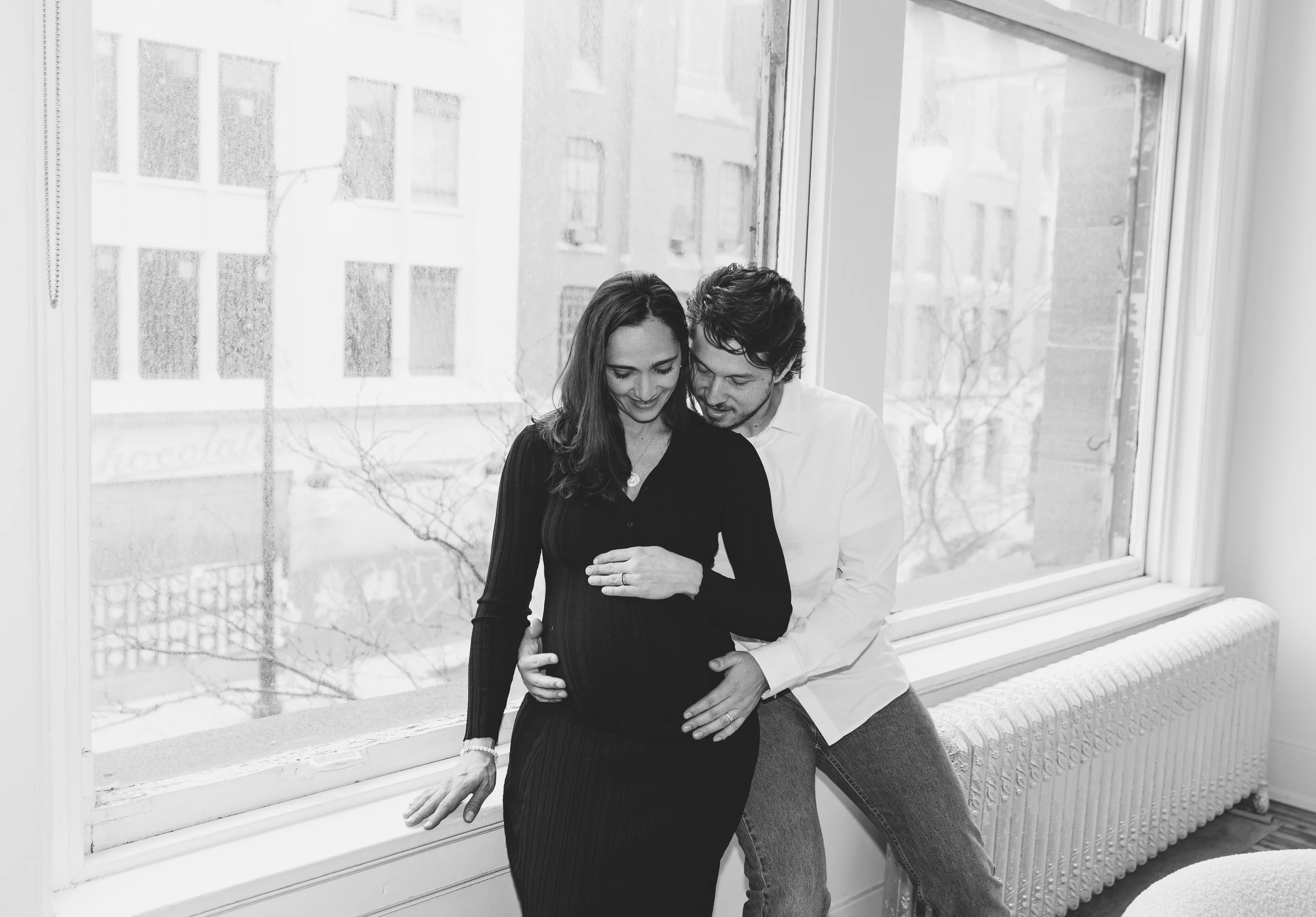 A pregnant woman and her partner standing by a large window, smiling and touching her belly.