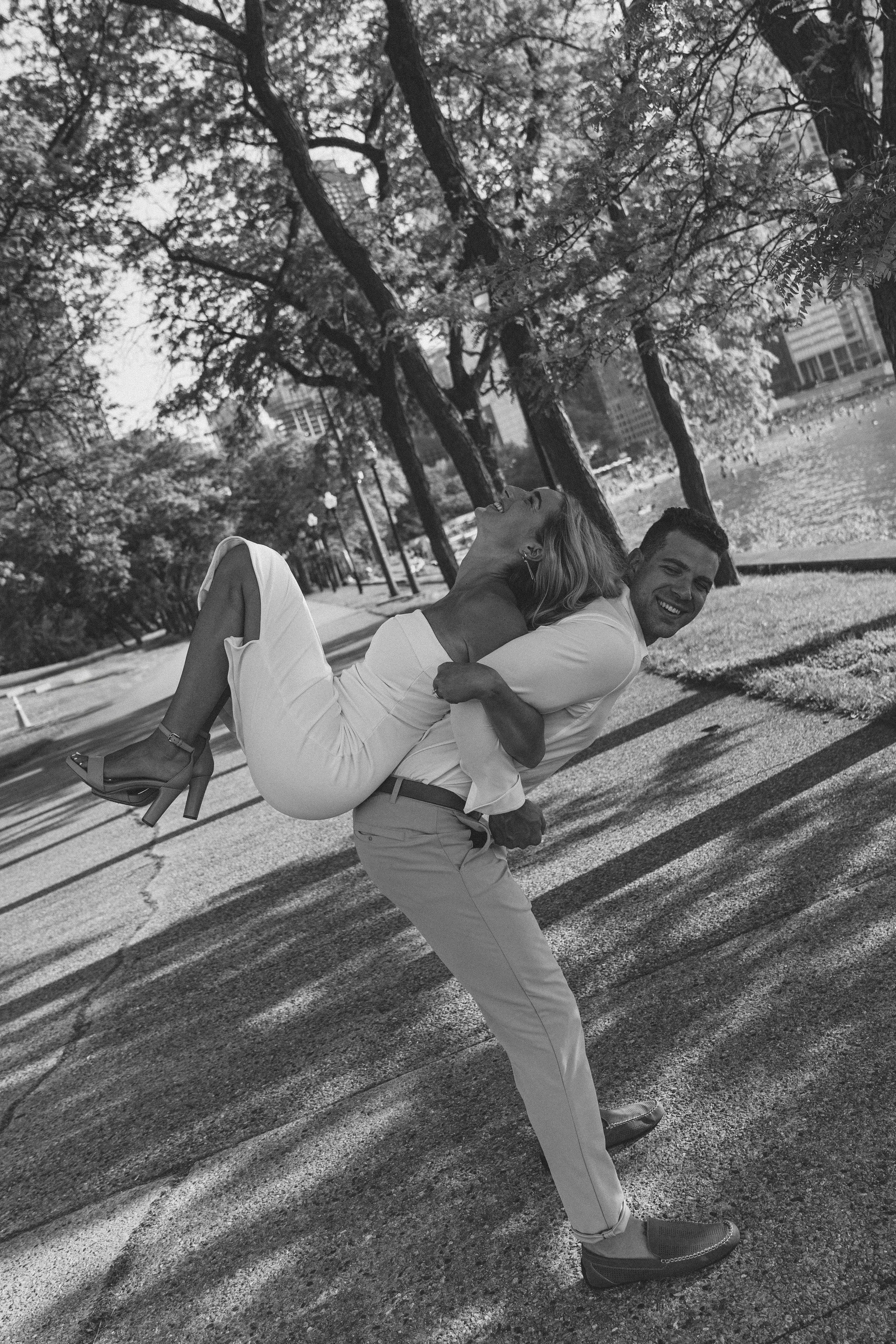A man in dress pants and a shirt is carrying a smiling woman in a dress and high heels in his arms, outdoors in a park with trees and a paved walkway in the background.