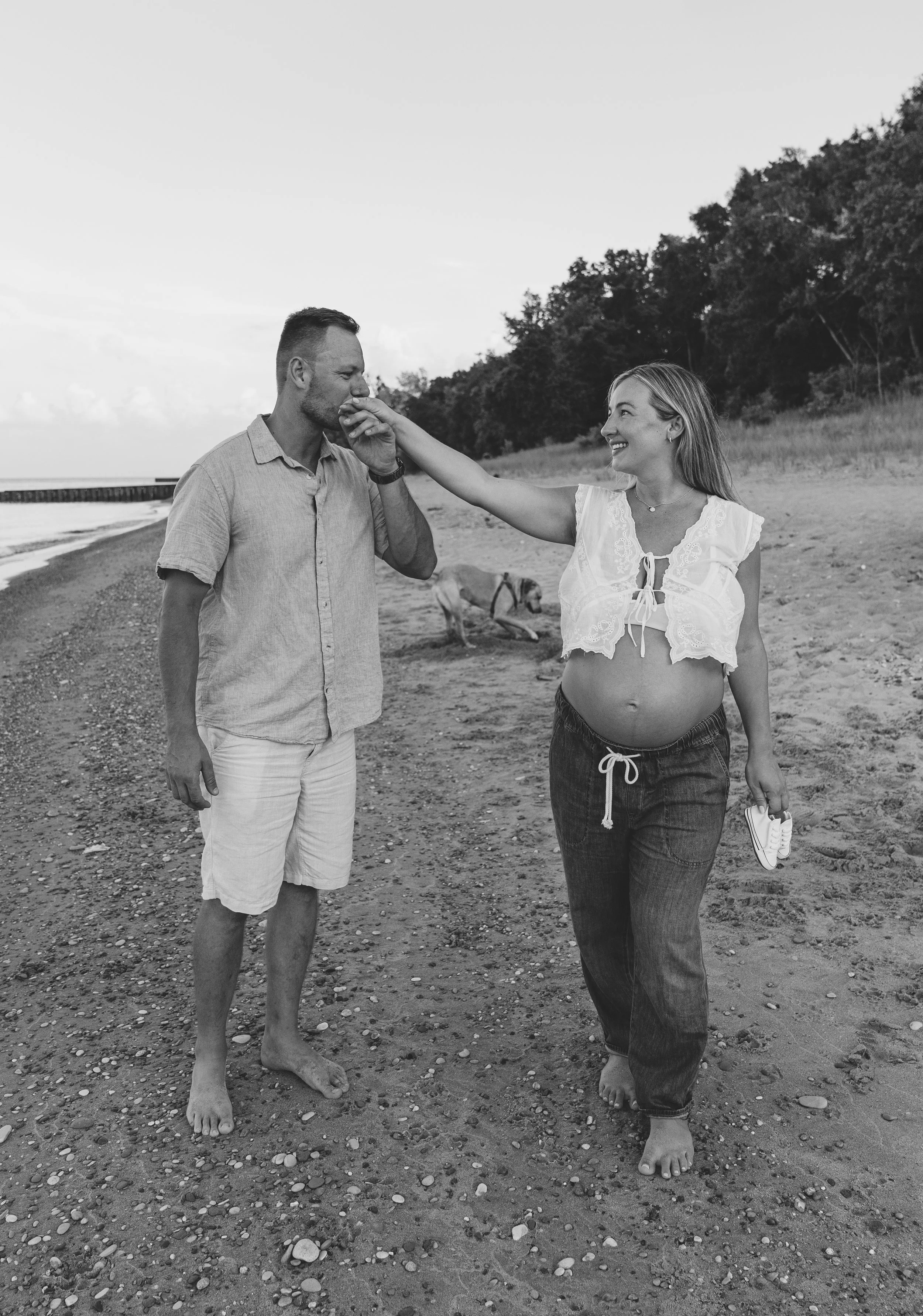 A pregnant woman on a beach is touching a man's face while smiling, as he covers his mouth with his hand. They are barefoot, and a dog is in the background near the shoreline. The scene is peaceful and affectionate.