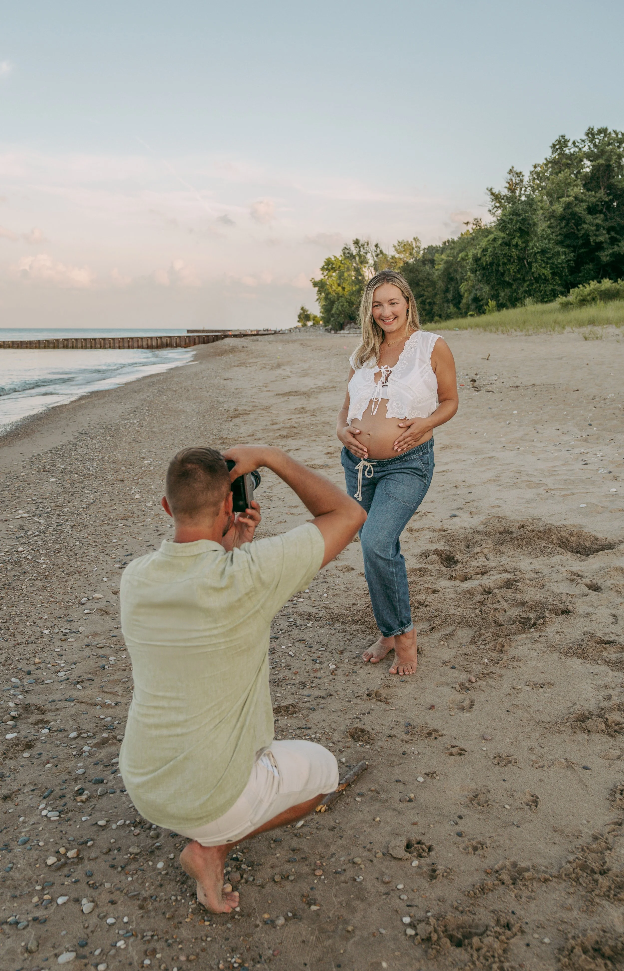 A woman in a white lace top and jeans is pregnant and stands on a sandy beach as a man kneels taking her photo, with trees and the ocean in the background.