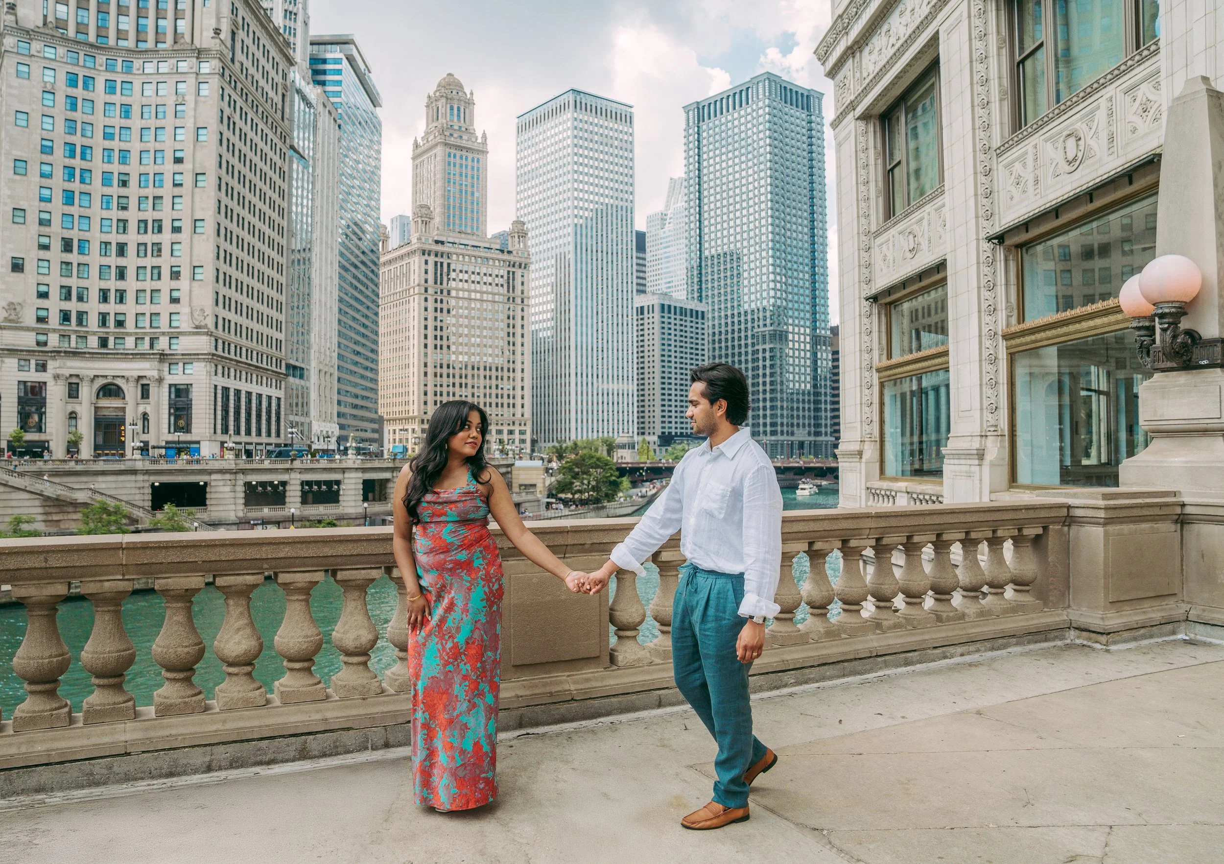 A man and woman holding hands on a bridge with a city skyline of tall buildings behind them.
