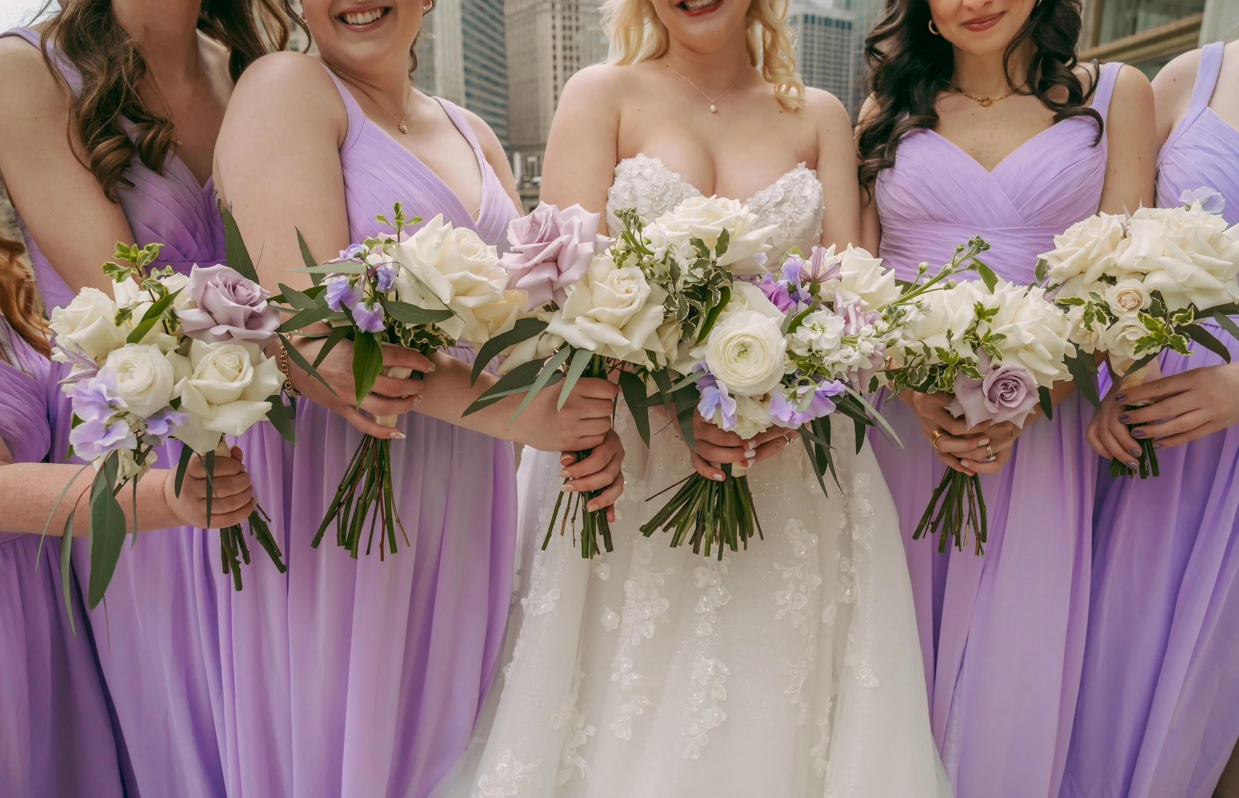 Close-up of a bride in a wedding dress holding a bouquet of white and purple flowers, surrounded by bridesmaids in lavender dresses also holding bouquets, with a cityscape in the background.