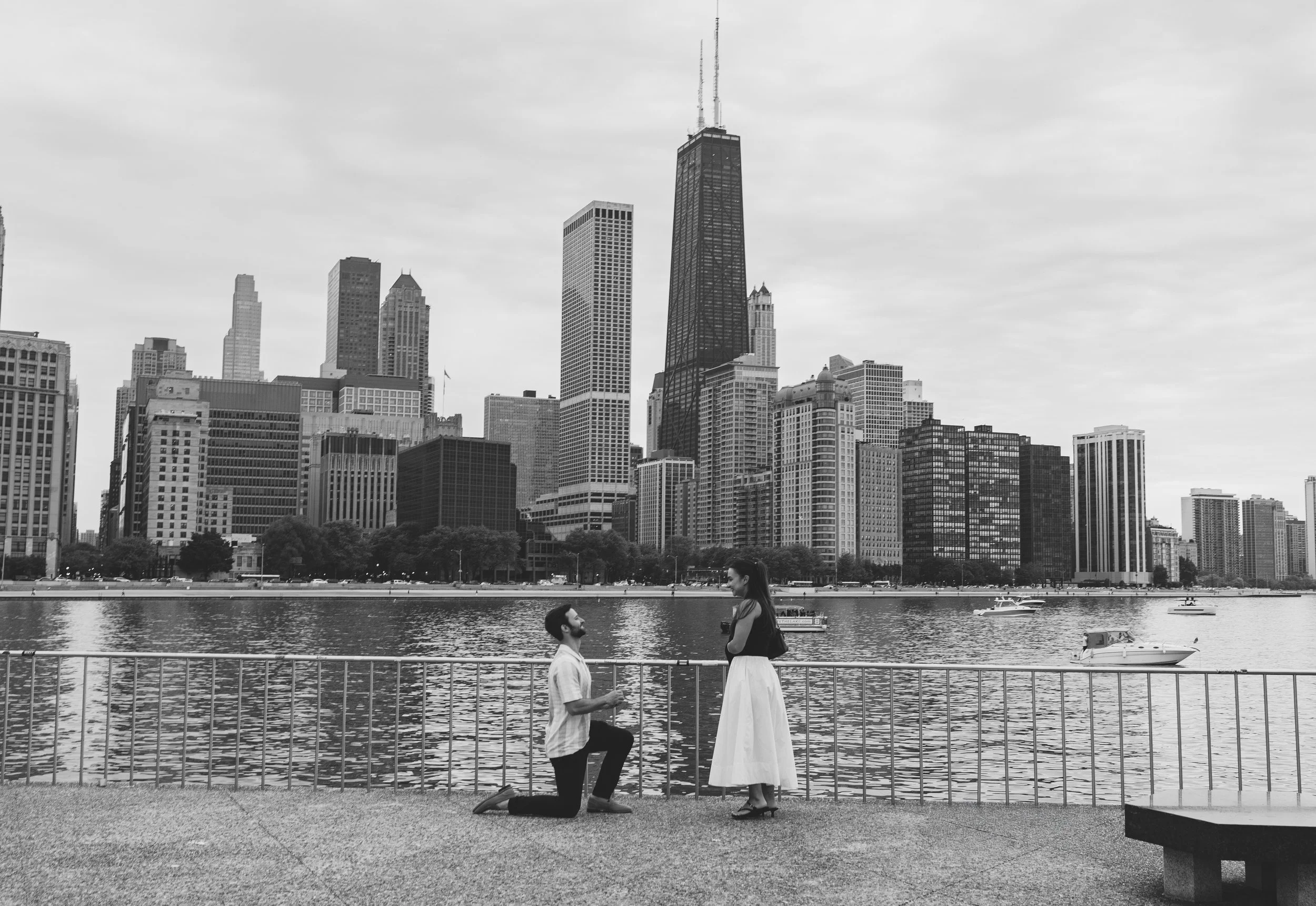 A man kneeling on one knee proposing to a woman by the water with a city skyline in the background.