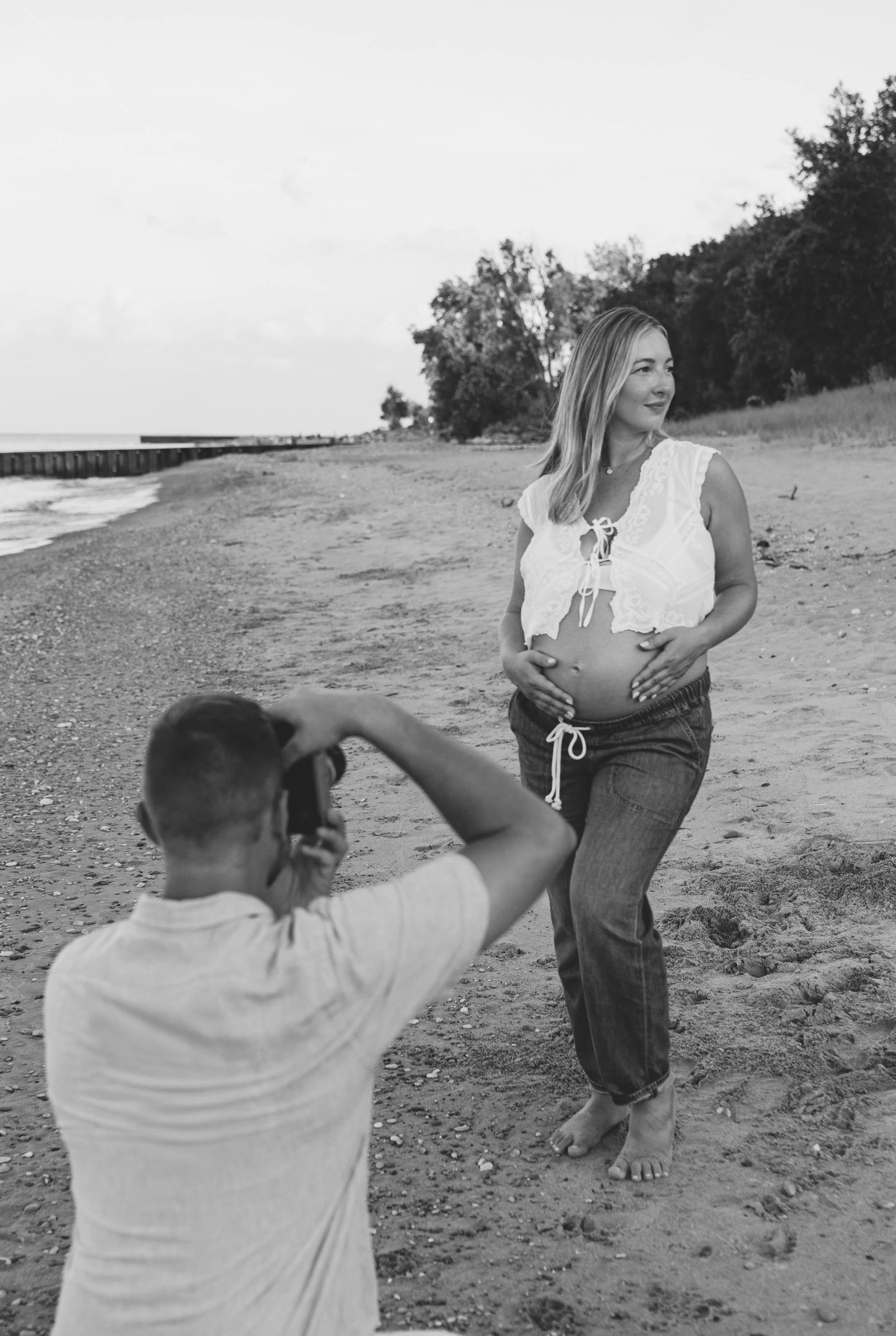 A pregnant woman in a white top and jeans standing on a beach, holding her belly, as a man takes her photo.