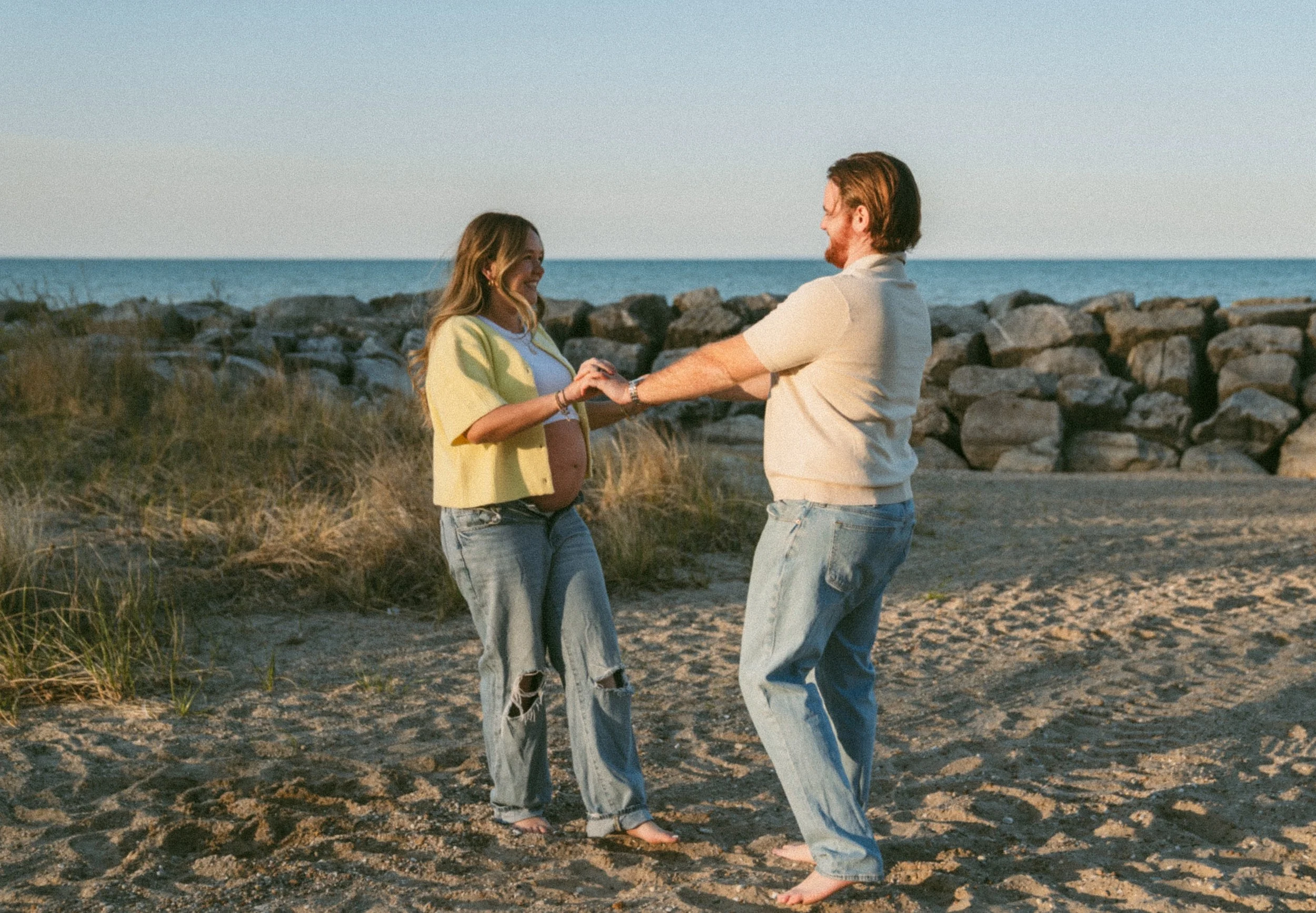 A couple dancing and holding hands on a sandy beach near rocks, with ocean and clear sky in the background.