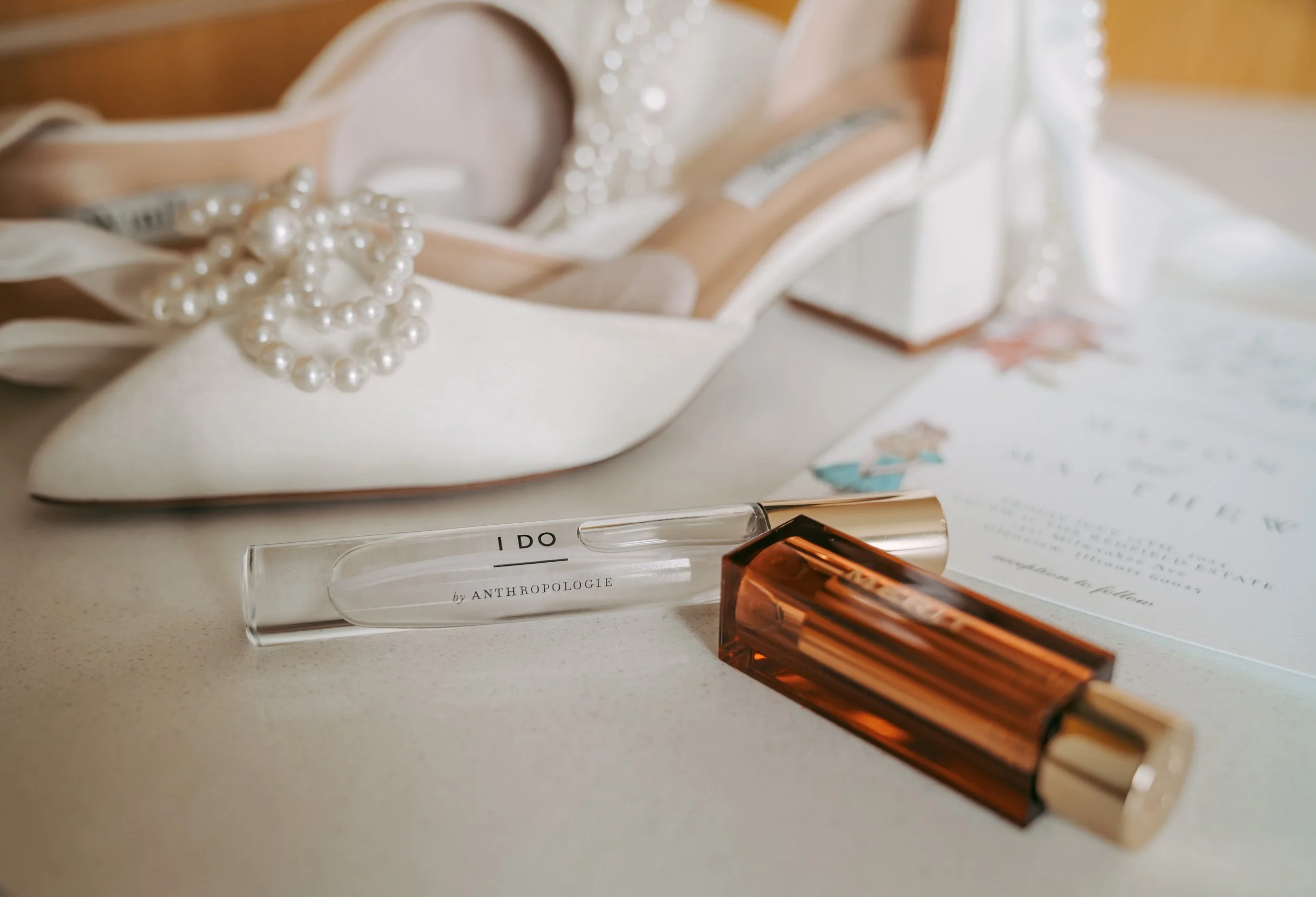 A pair of white high heel shoes with pearl embellishments, a vial of perfume labeled 'I DO' by Anthropologie, a rectangular amber-colored perfume bottle, and a handwritten note on decorative paper.