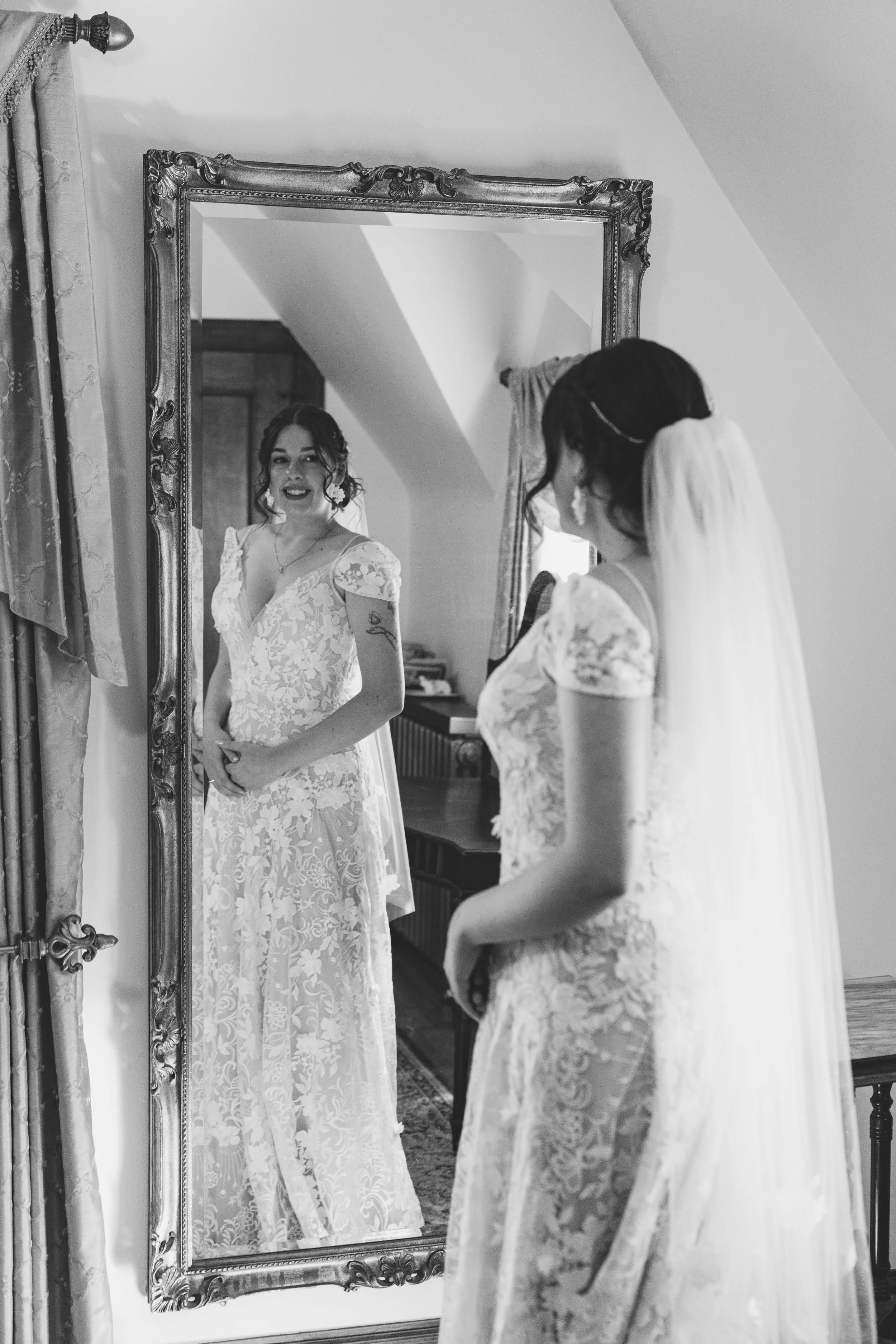 A bride in a lace wedding dress looking at herself in a large ornate mirror, smiling, with her reflection showing her face and upper body.