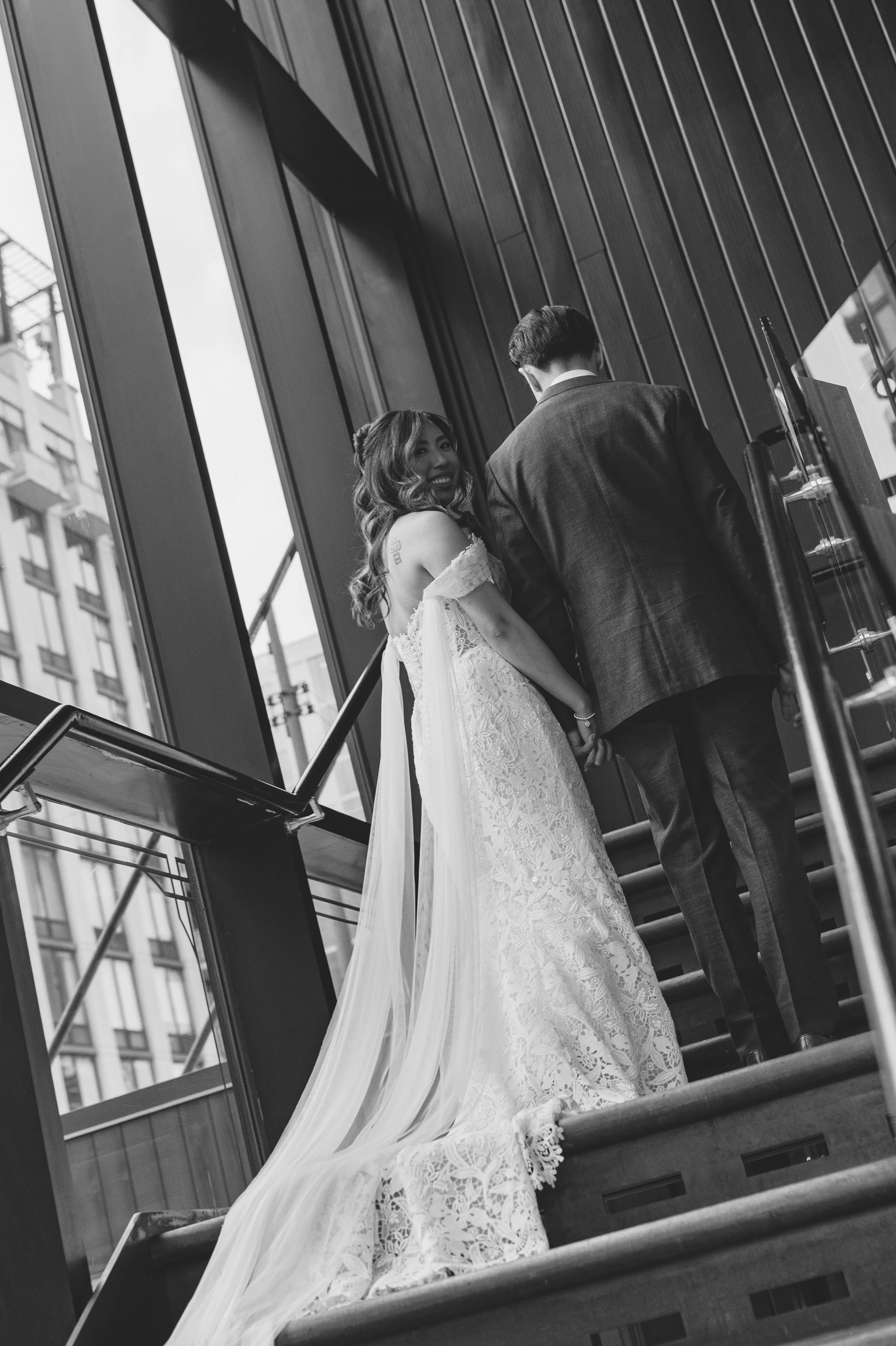 A bride and groom holding hands on a staircase, taken from behind, with tall glass windows and city buildings in the background, in black and white.