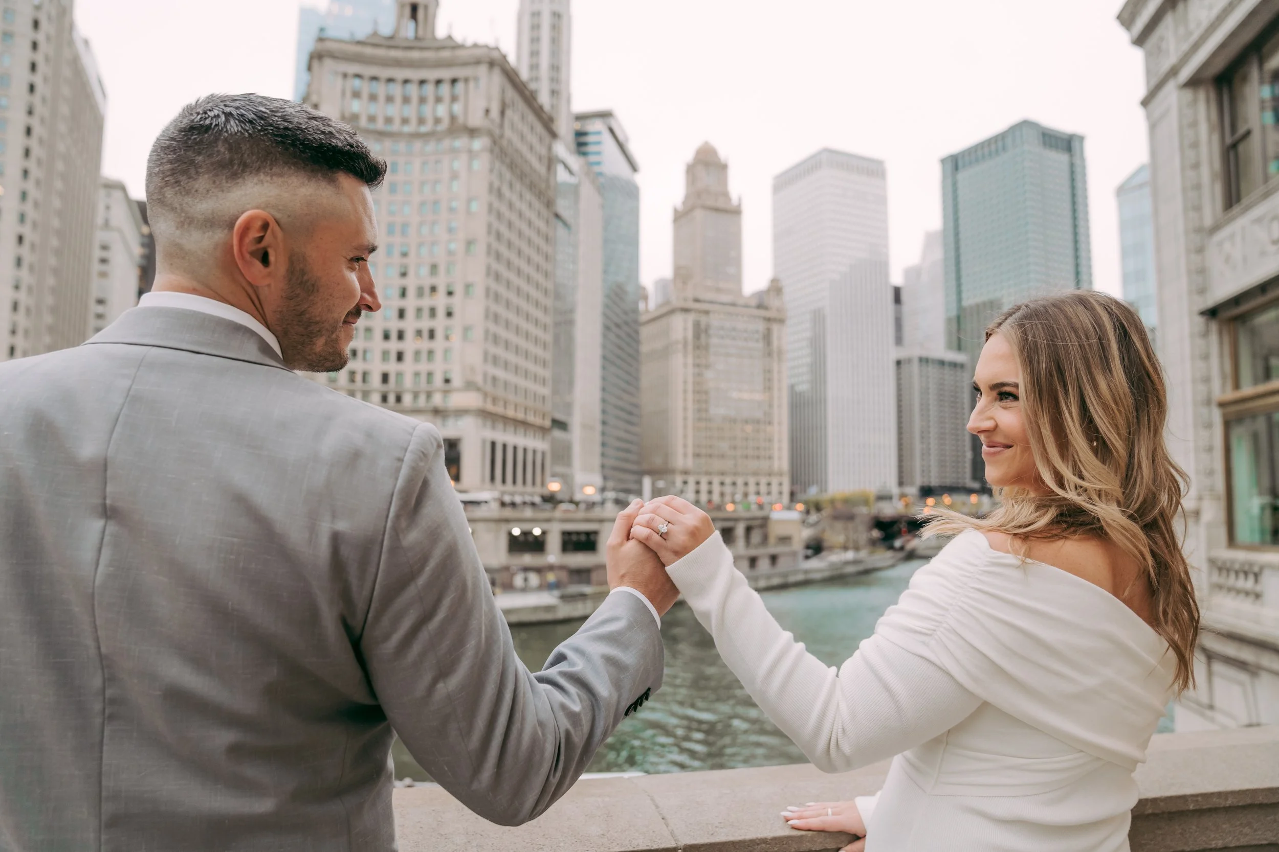A man in a light gray suit holding hands with a woman in a white off-shoulder dress, both standing on a bridge overlooking water with a city skyline in the background, sharing a romantic moment.