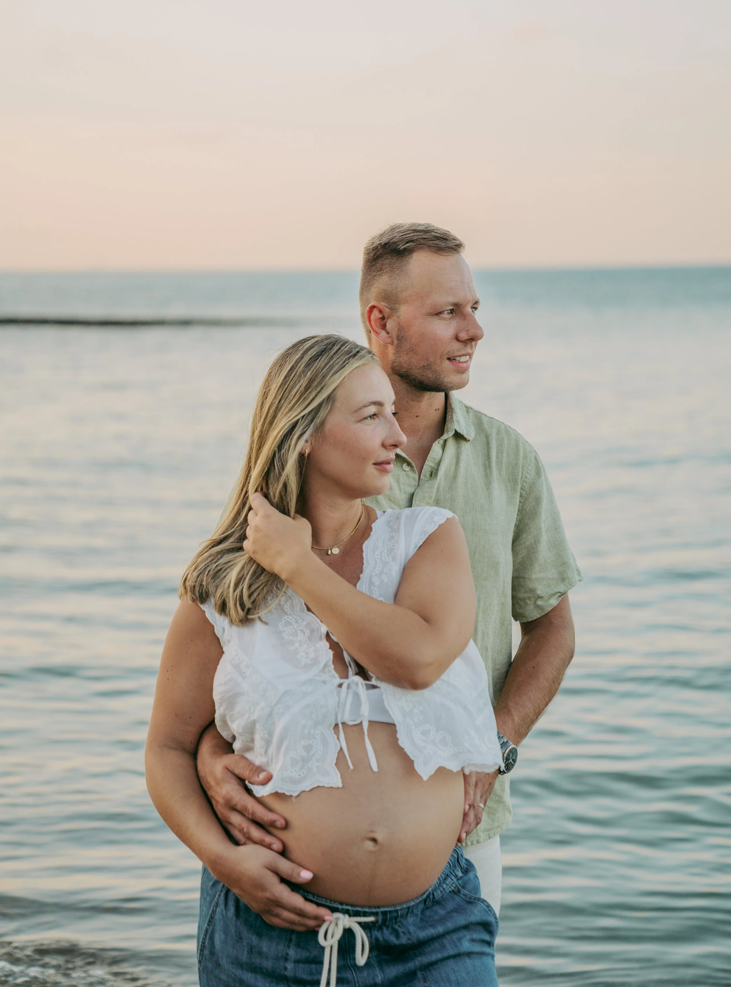 A pregnant woman and a man stand on a beach at sunset, with the woman gently touching her baby bump and the man's hand resting on her stomach.