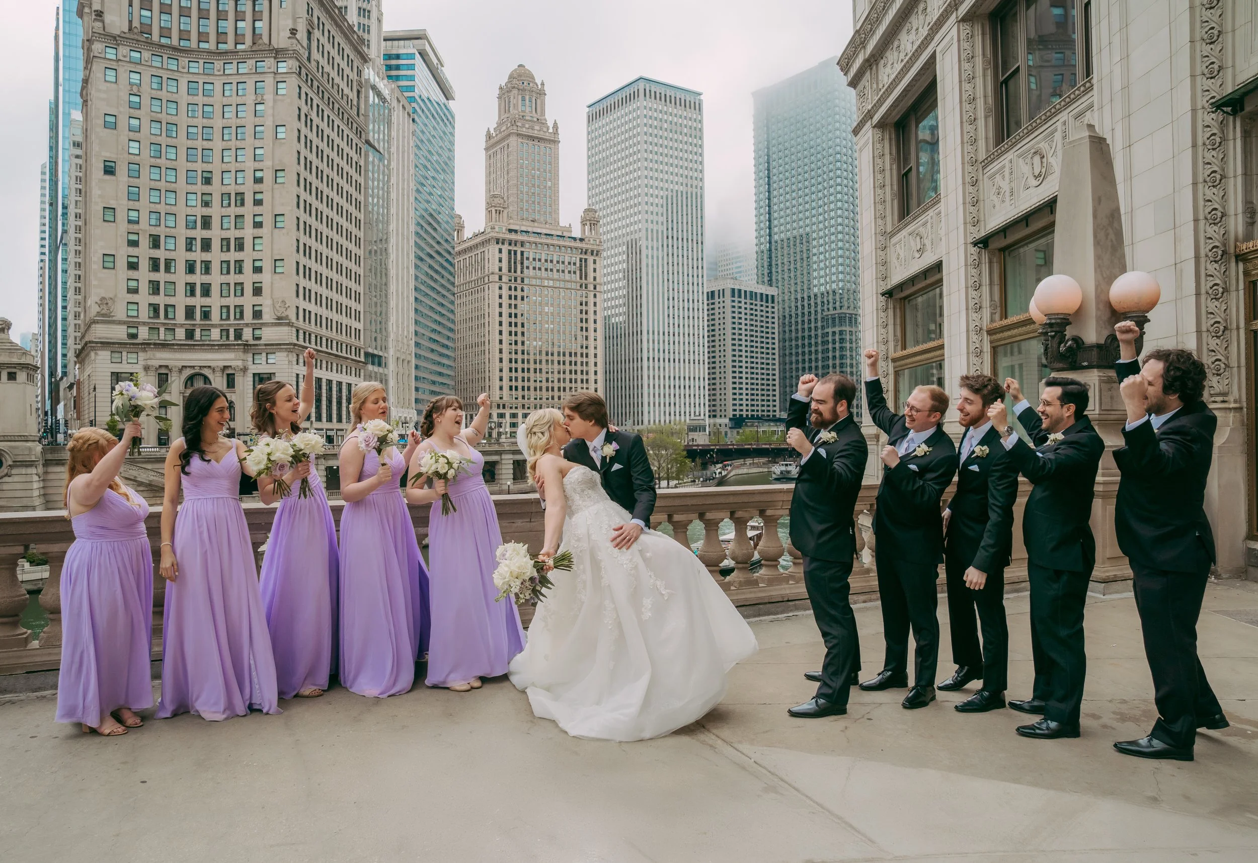 A wedding party in wedding attire, with the bride and groom kissing, surrounded by bridesmaids in lavender dresses and groomsmen in black suits, celebrating on a rooftop in an urban city with tall buildings.