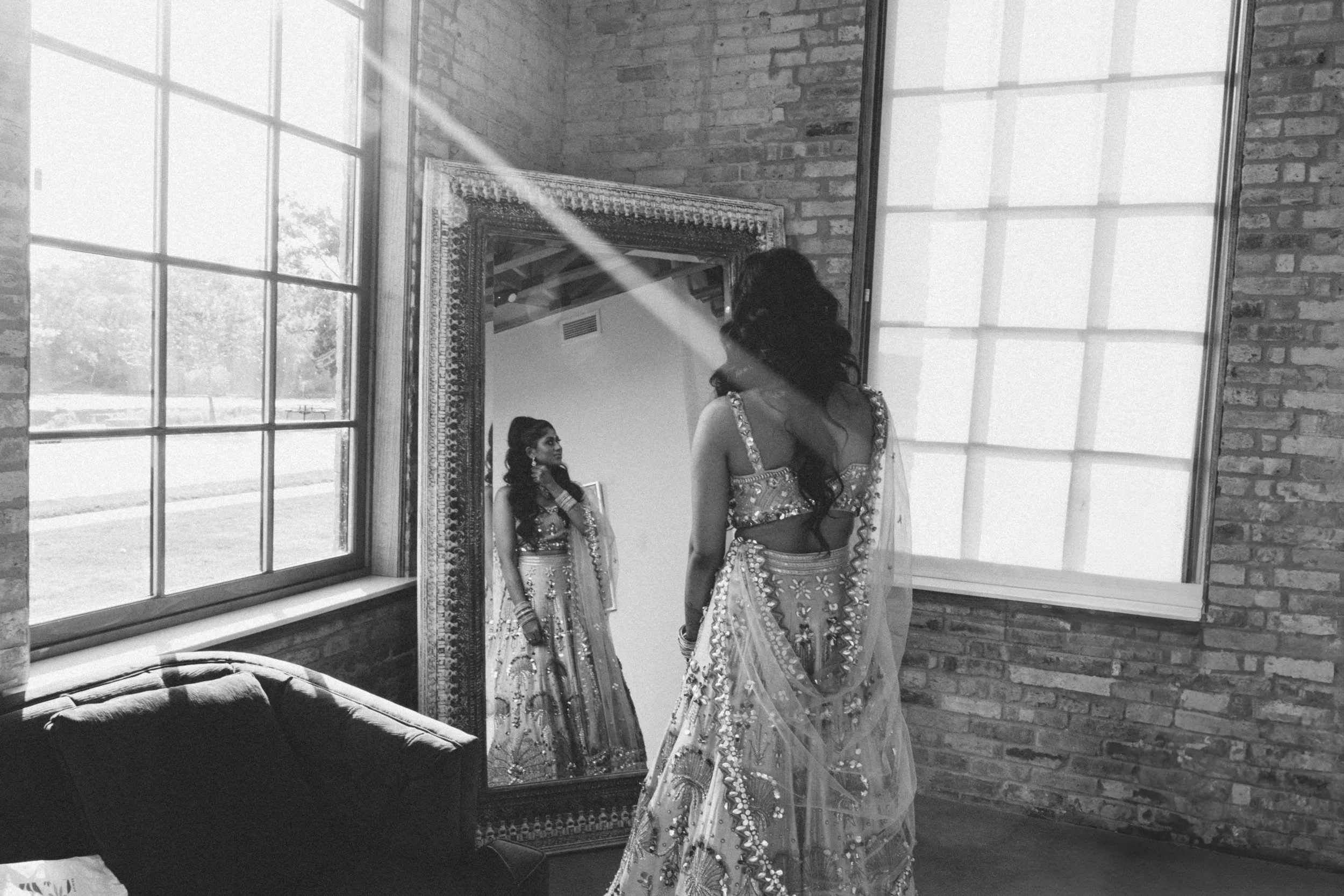 A woman dressed in traditional Indian attire looking at herself in a tall mirror in a room with brick walls and large windows.