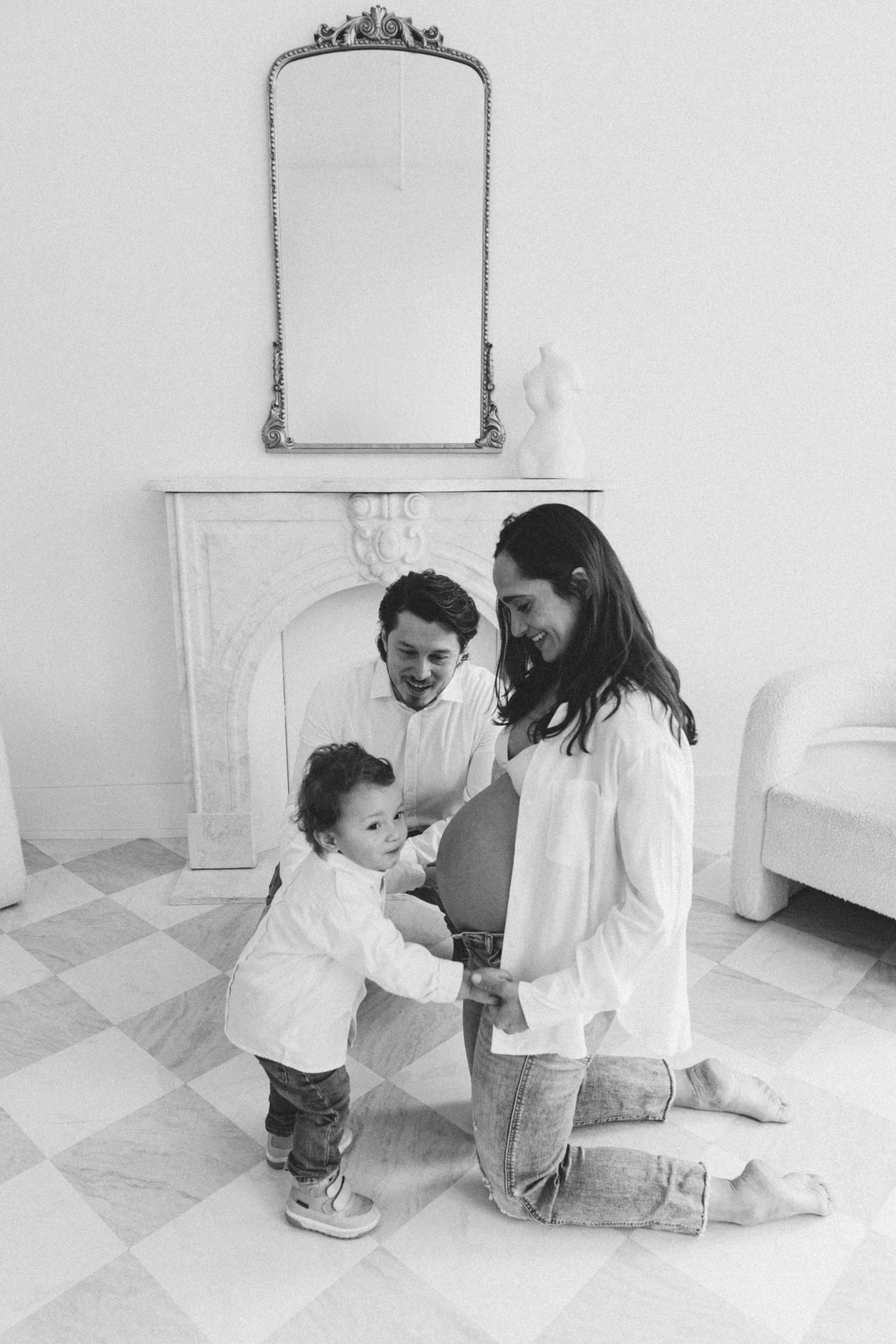 A pregnant woman kneels on the floor, holding her belly, with a child and a man. The man and child hold her belly, smiling. The room has a fireplace, a mirror, and a sculpture on the mantel.