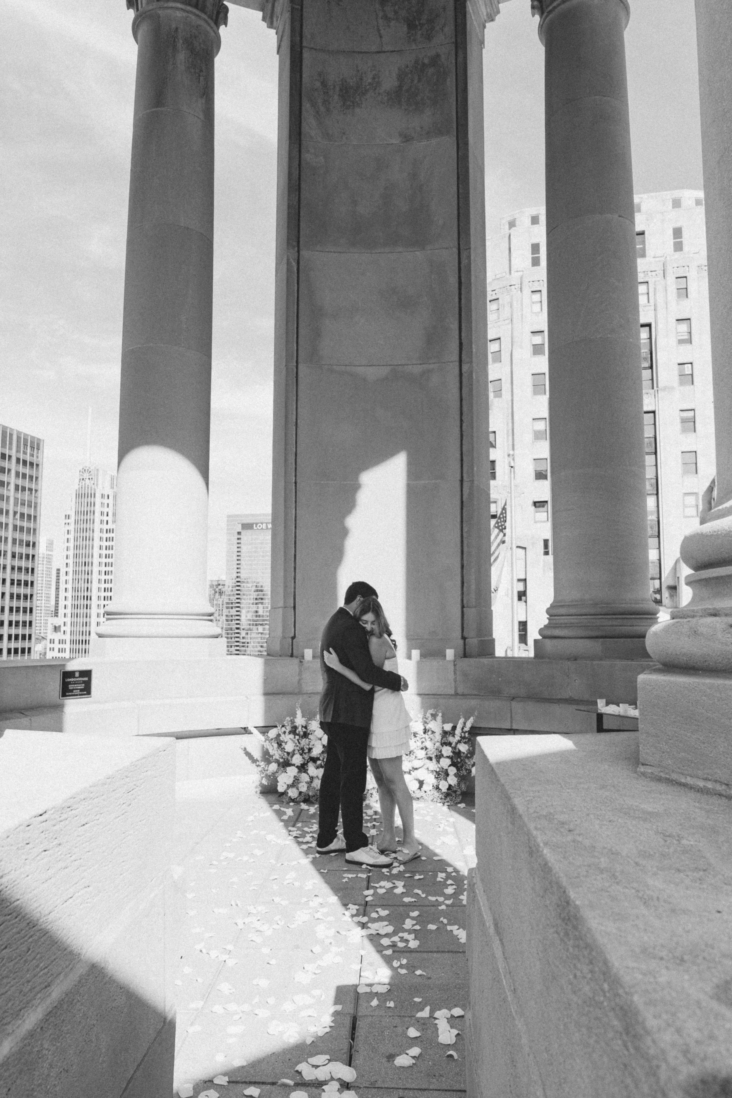 A couple hugging at a wedding ceremony on a terrace, with flower petals on the ground and a cityscape in the background, black and white photo.
